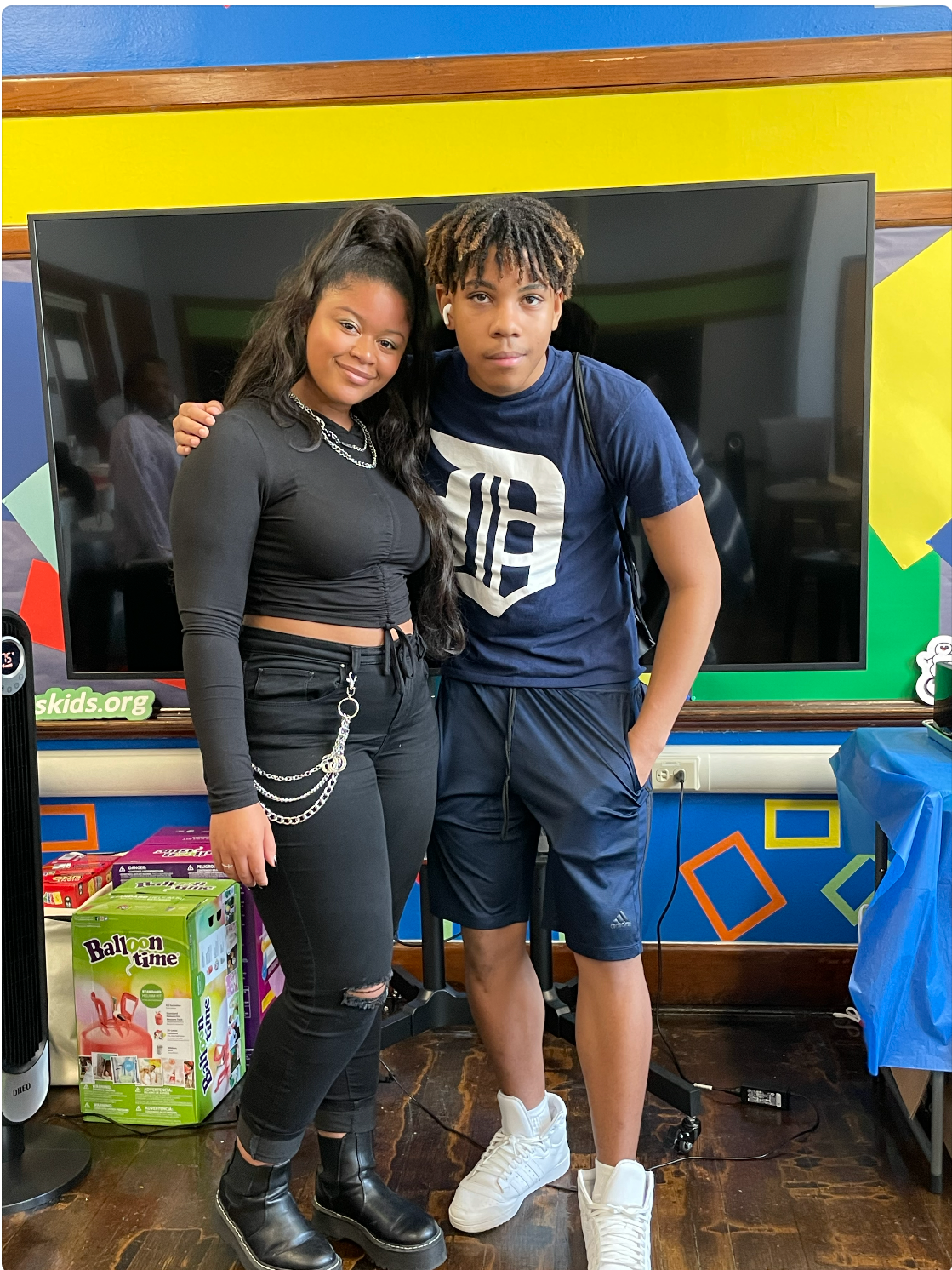 Two teenagers, a girl and a boy, posing together indoors in front of a large screen. The girl wears a black long-sleeve crop top, ripped black jeans, black boots, and has long dark hair. The boy wears a blue t-shirt with a large white logo, blue athletic shorts, and white sneakers, with dreadlocks and AirPods. They are standing close, with the girl smiling at the camera and the boy looking serious.