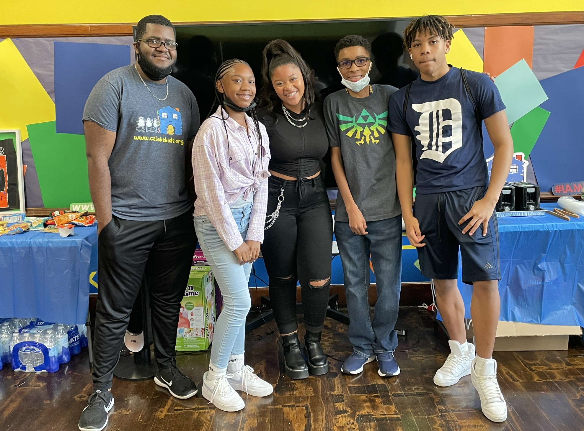 Group of six diverse people standing together in a room with colorful decorations and snack tables behind them.