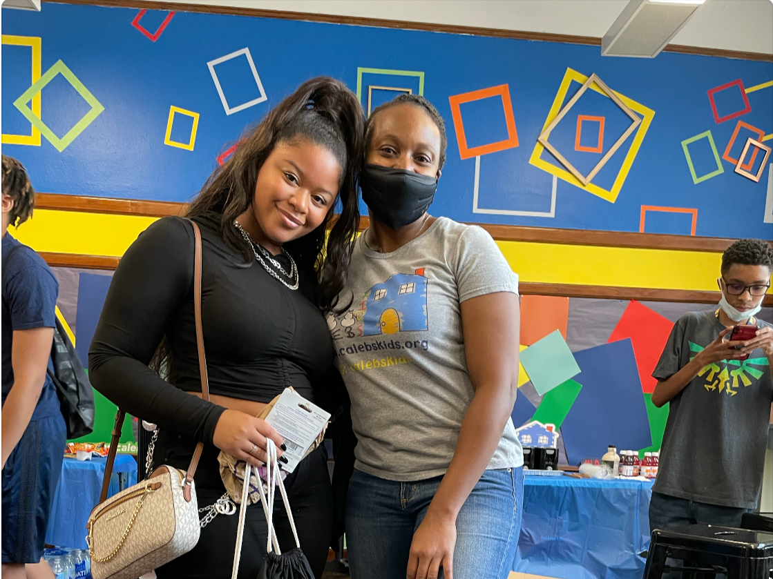 Two women standing close together, smiling at the camera, at a colorful indoor event. The woman on the left has long black hair and is wearing a black top and a purse. The woman on the right has short hair, wearing a gray T-shirt with a logo and a black face mask. In the background, a boy is looking at his phone, wearing glasses and a face mask, with a table of items behind him.
