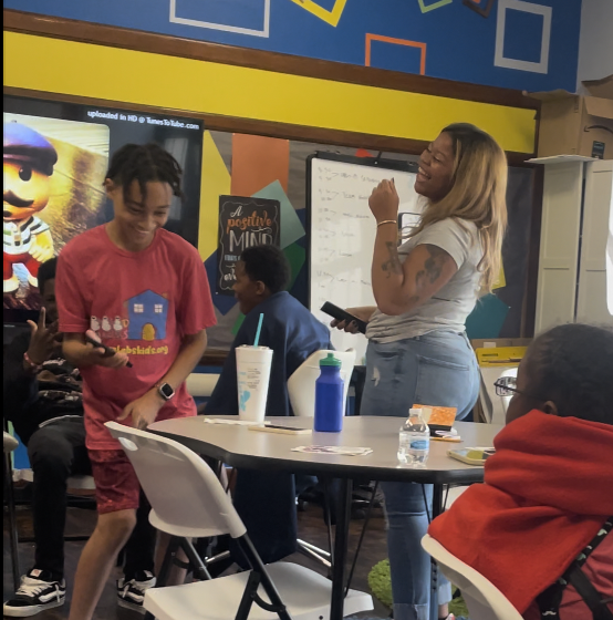 A classroom with students and a teacher. One student is smiling and wearing a pink shirt, standing beside another student in a blue shirt. The teacher, with long hair and tattoos, is holding a phone and appears to be speaking or explaining something. There is a whiteboard behind her, and the classroom has colorful decorations and a large digital screen.