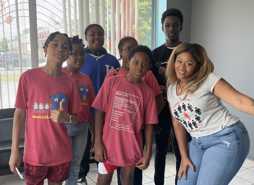 Group of seven children and one adult woman standing indoors near windows with vertical blinds, smiling for the camera.