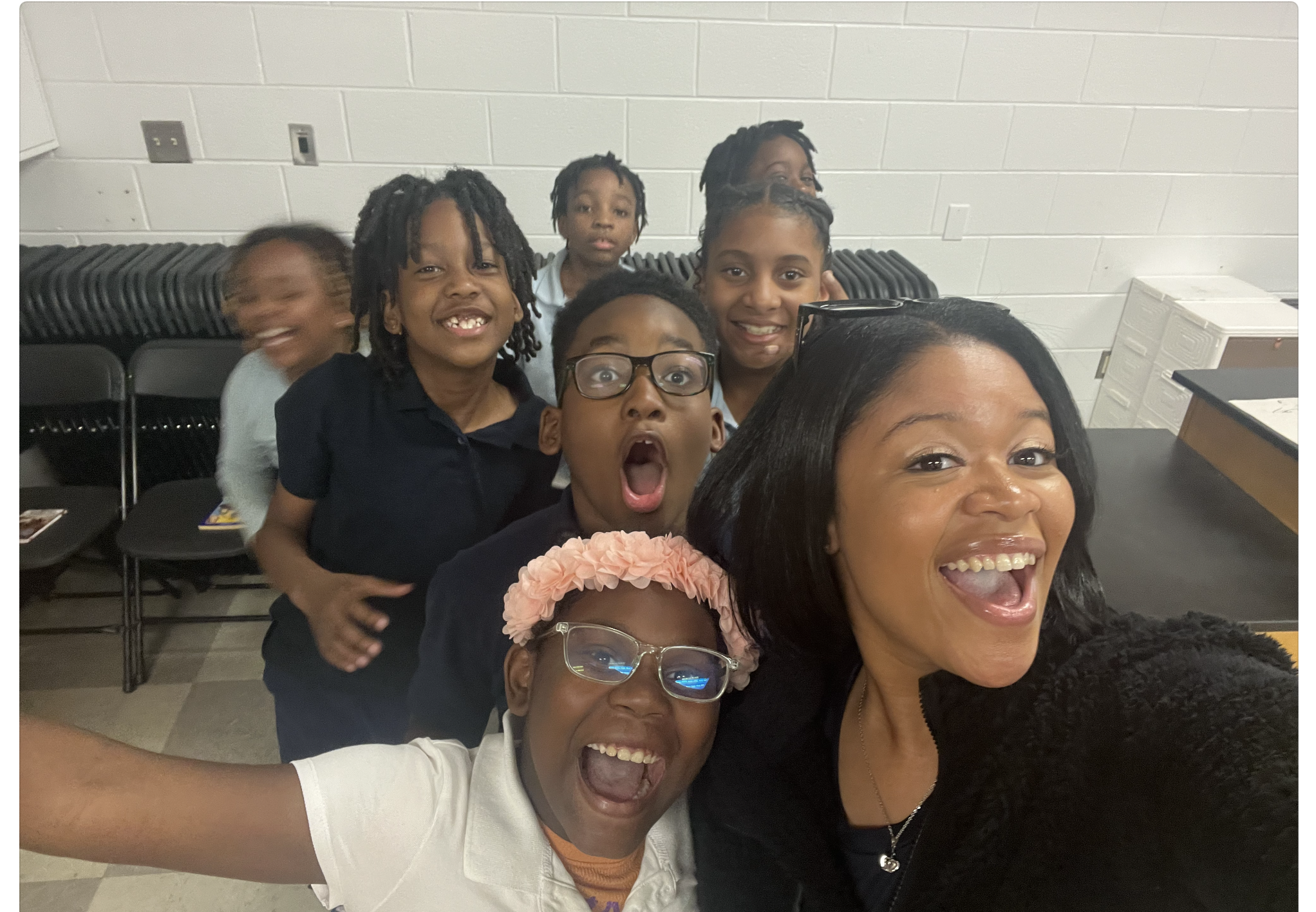 A woman taking a selfie with seven children in a classroom setting, all smiling and expressing excitement.