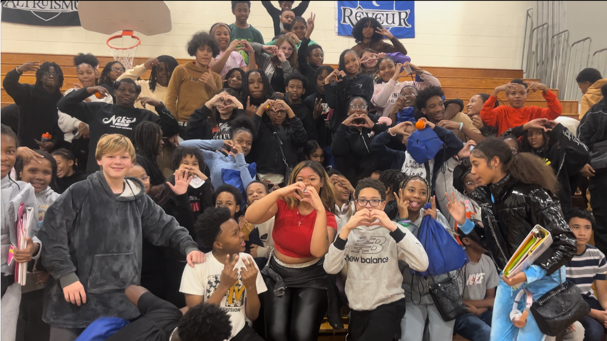 A large group of children and teenagers posing together in a gymnasium with wooden bleachers. Some are making heart shapes with their hands, while others are smiling and waving. The group appears to be at school or a community event.