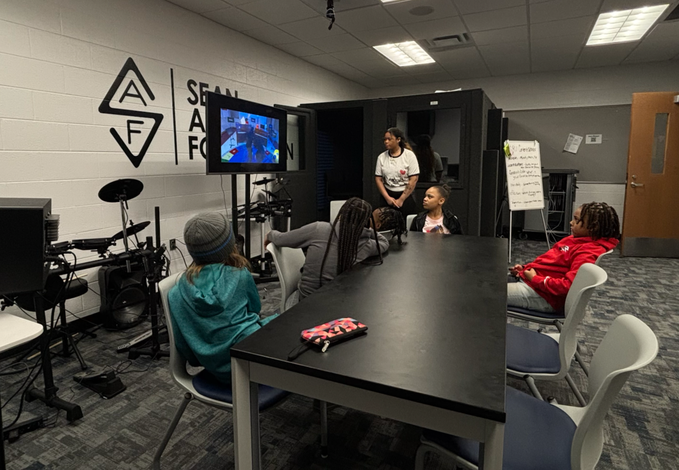 Children watching a presentation or video on a monitor in a classroom or workshop setting, with an instructor present, in a room with AV equipment and a wall sign with a logo