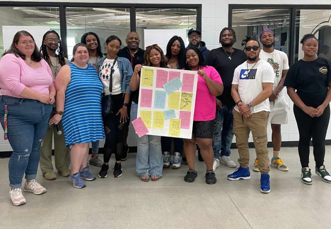 Group of diverse individuals standing in front of a window, holding a large poster with colorful notes and drawings.