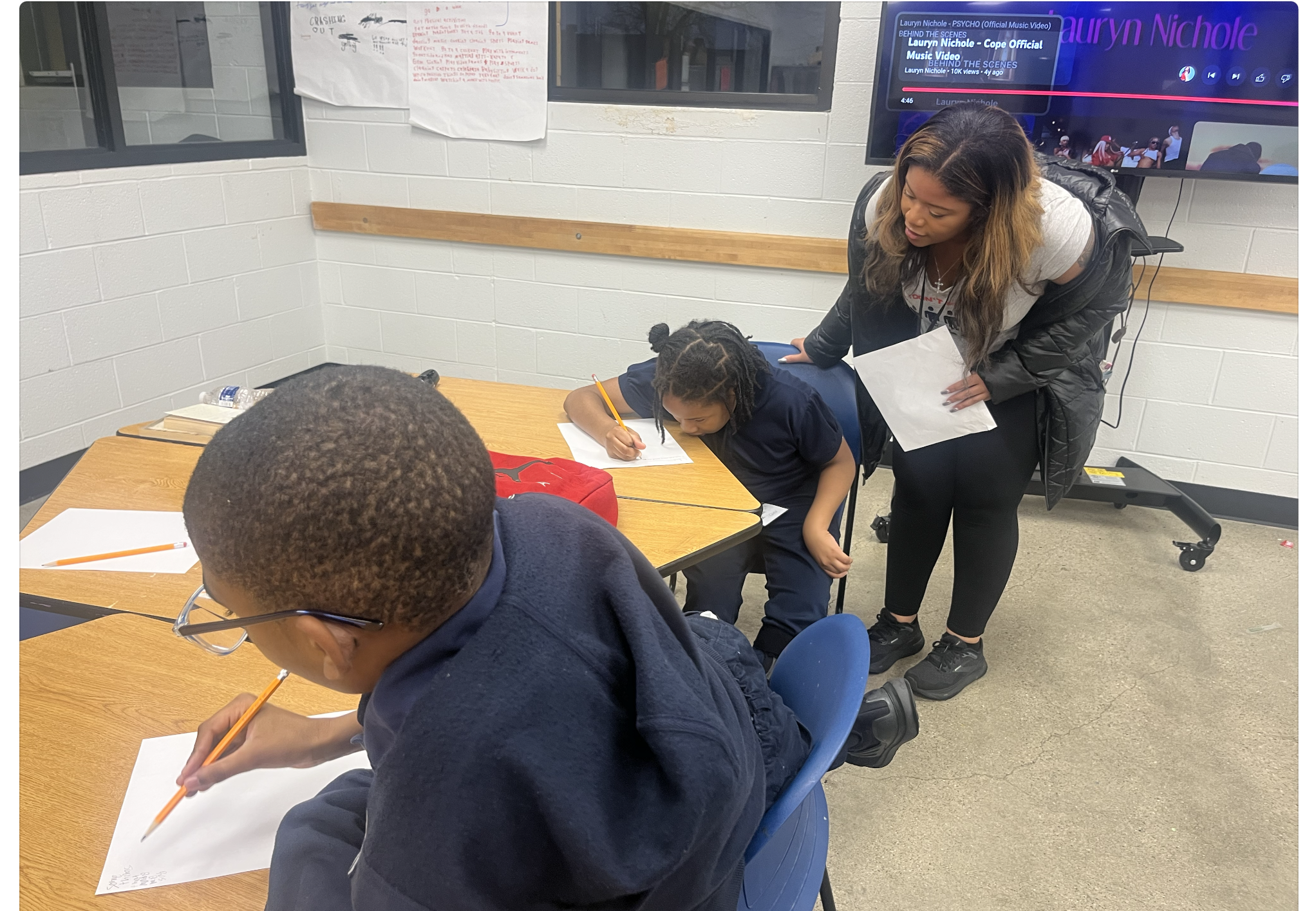 A classroom scene with two children seated at a table, each writing on sheets of paper with pencils, and a woman standing beside them supervising or assisting. The classroom has light gray brick walls, windows, and a large monitor displaying a YouTube video featuring Lauryn Nichole.
