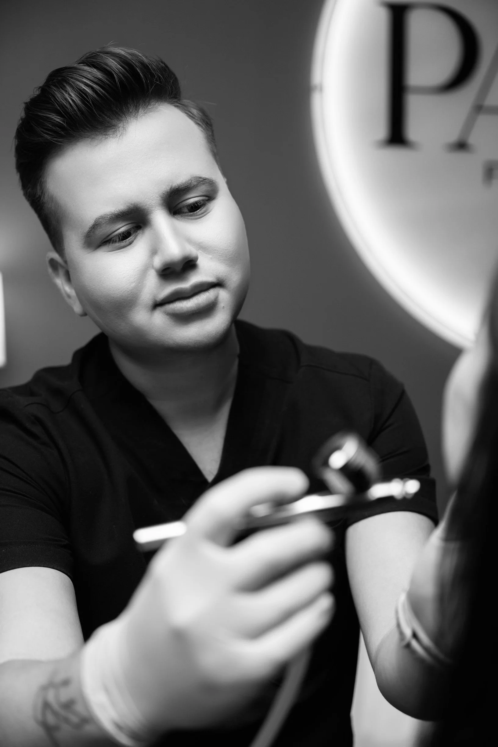 A young person with short hair in black scrubs, wearing gloves, appears to be inspecting or preparing a tattoo machine in a tattoo studio.