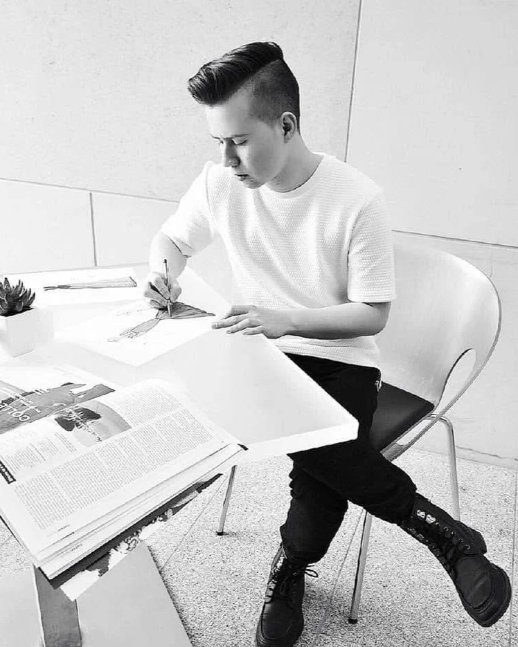 A young man with a mohawk hairstyle sitting at a table, drawing or writing on a piece of paper with a pen. There are magazines or newspapers open on the table, and a small potted plant is nearby. The photo is in black and white.