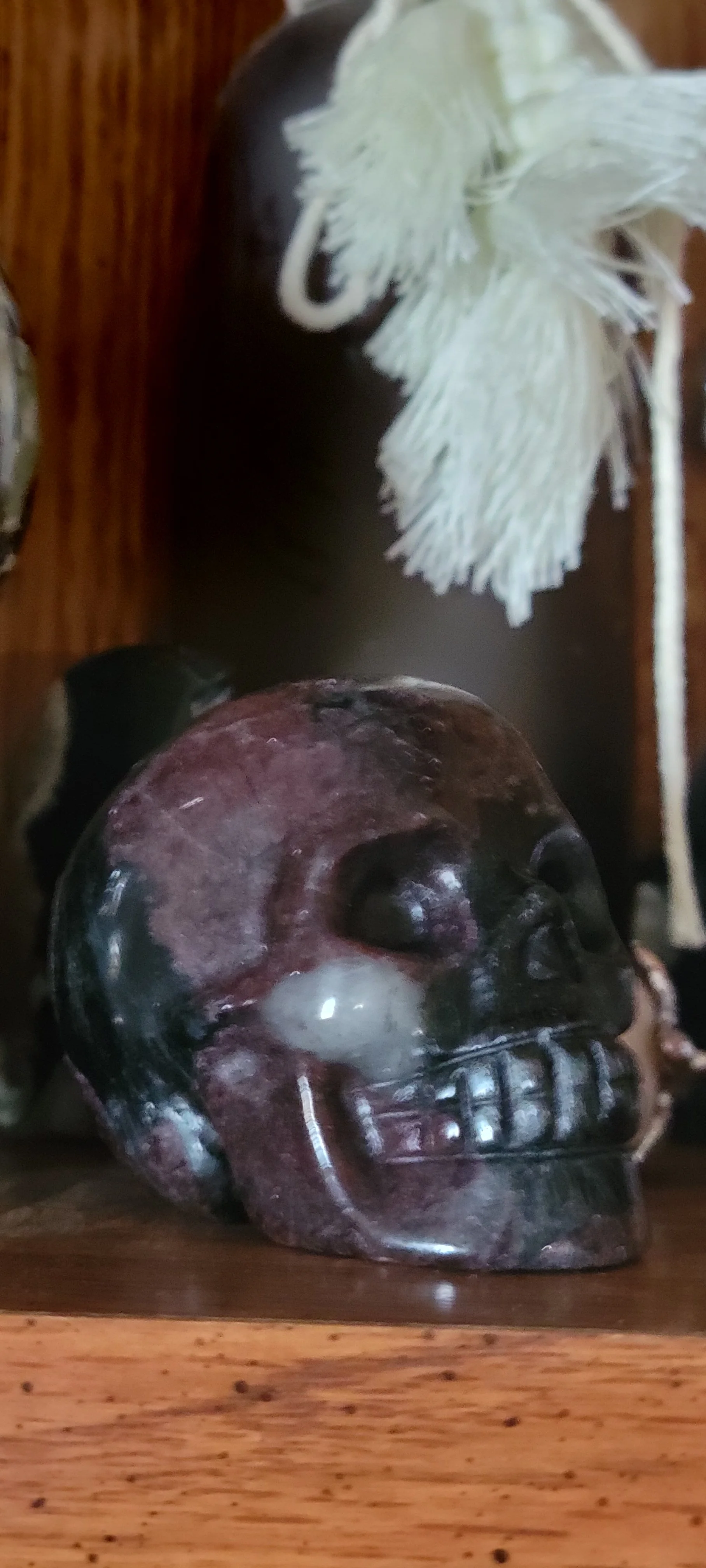 A decorative skull sculpture made of multicolored stone, placed on a wooden surface with a dried white flower in the background.