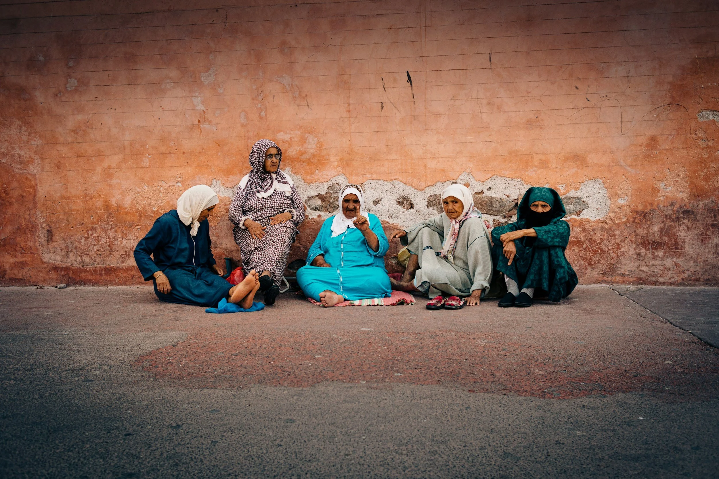 Five Moroccan women wearing colorful headscarves gathered in conversation against a traditional pink wall in Morocco.