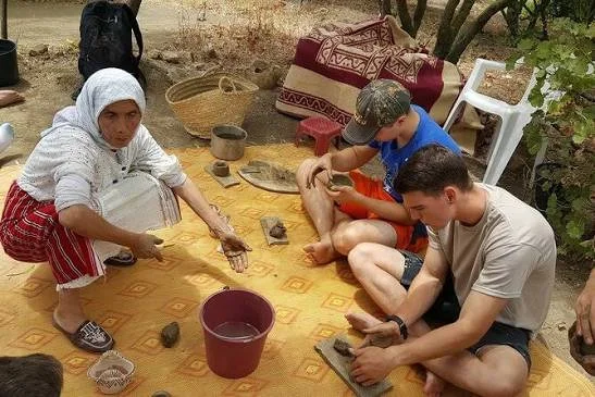 A woman and two men sitting on a yellow patterned mat outdoors, engaged in craft-making with clay or pottery; the woman is holding a clay object, and the men are shaping clay with their hands, surrounded by pottery tools and baskets.