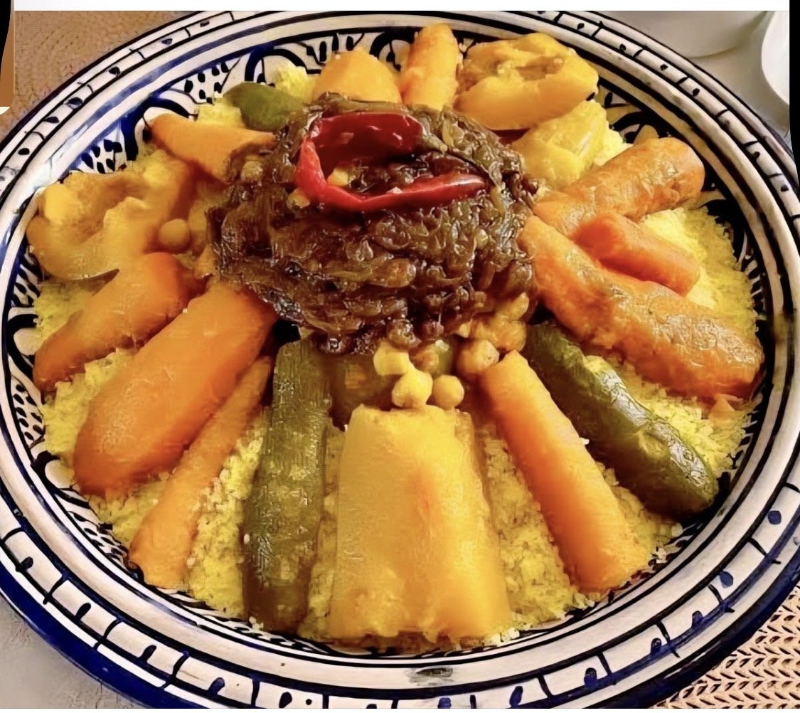 A colorful dish of cooked vegetables including carrots, zucchini, yellow squash, and eggplant, topped with a generous portion of cooked greens garnished with tomato slices, served over yellow couscous in a decorative bowl.