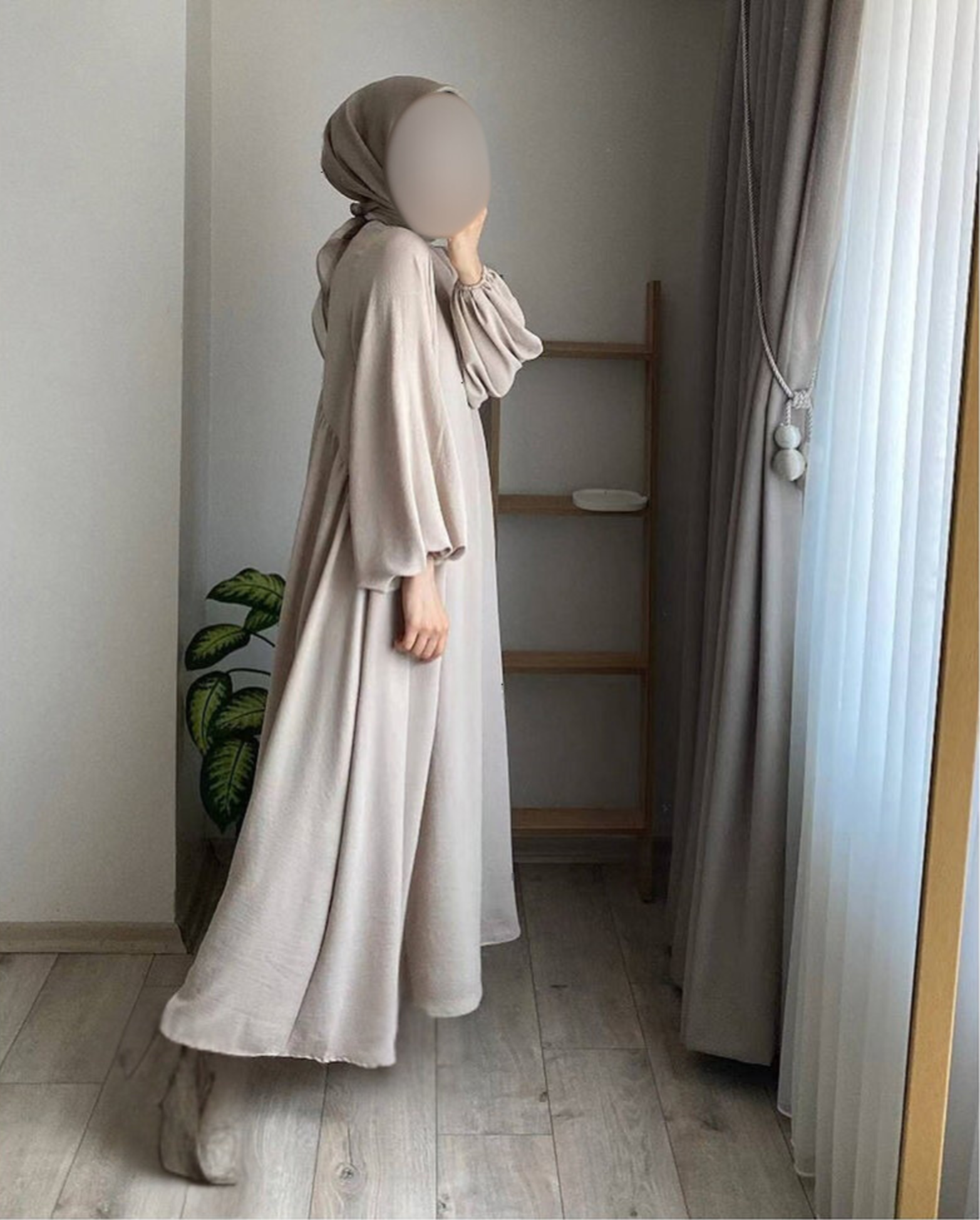 Person wearing a beige dress and a headscarf, standing indoors near a window with curtains, with a wooden shelf and a potted plant nearby.