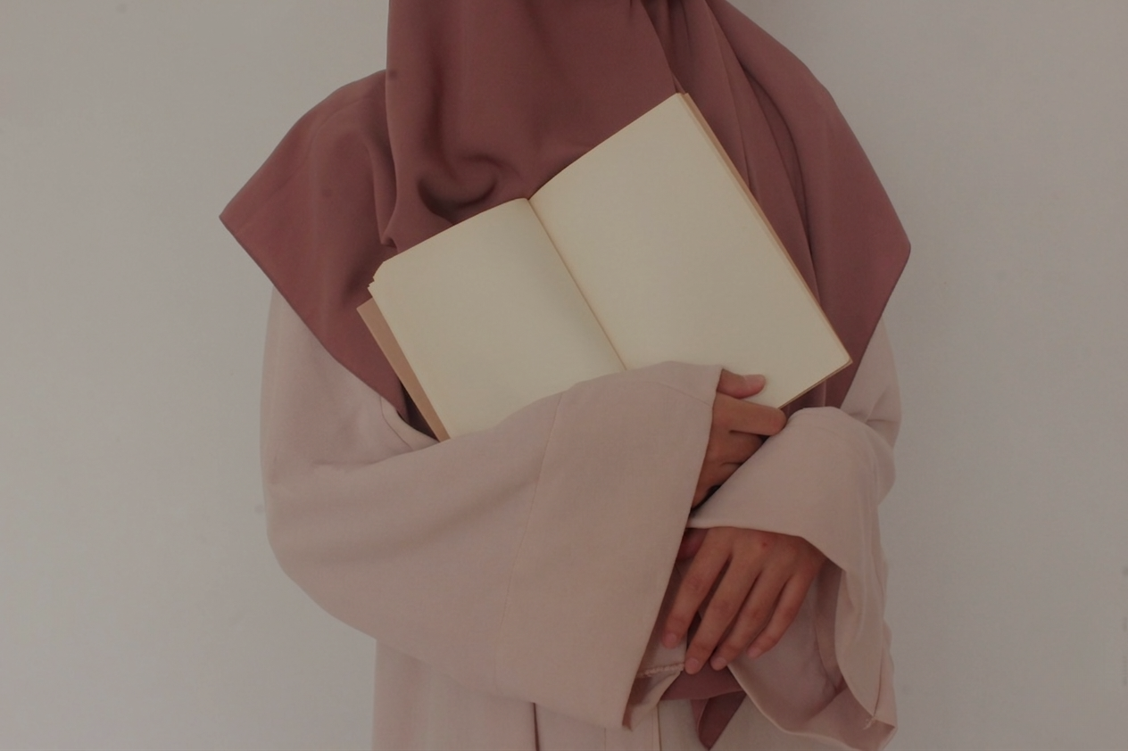 Person in a pink hijab holding an open blank notebook with both hands.