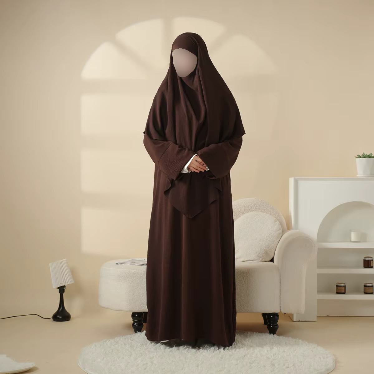 A person wearing a brown hijab and dress standing in a living room with beige walls and minimal furniture, including a white sofa, a small side table, a modern lamp, and a plant.