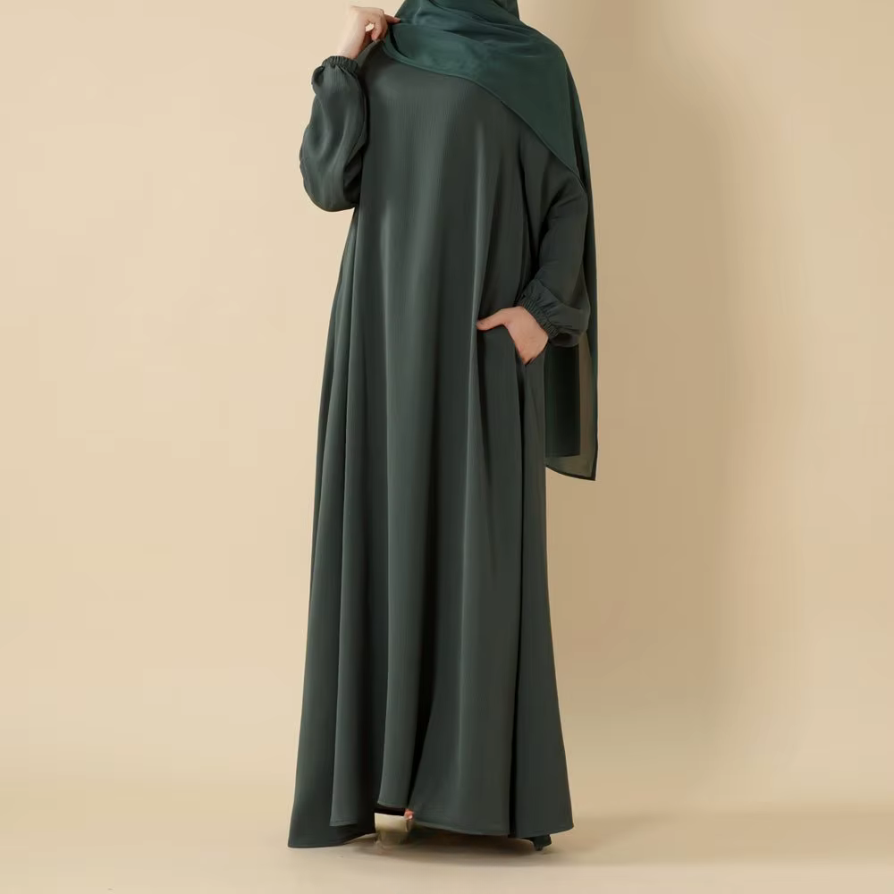 A woman wearing a dark green, flowing abaya with long sleeves and a matching headscarf, standing against a beige background.
