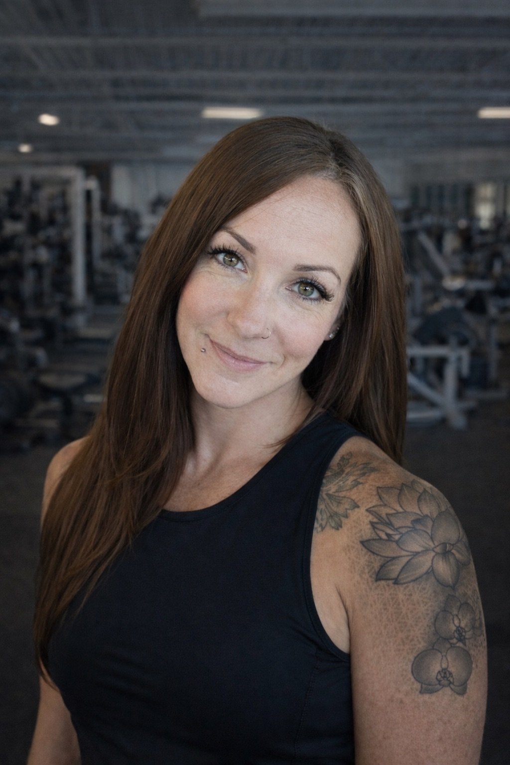 A woman with long brown hair and tattoos on her arm, smiling at the camera in a gym.