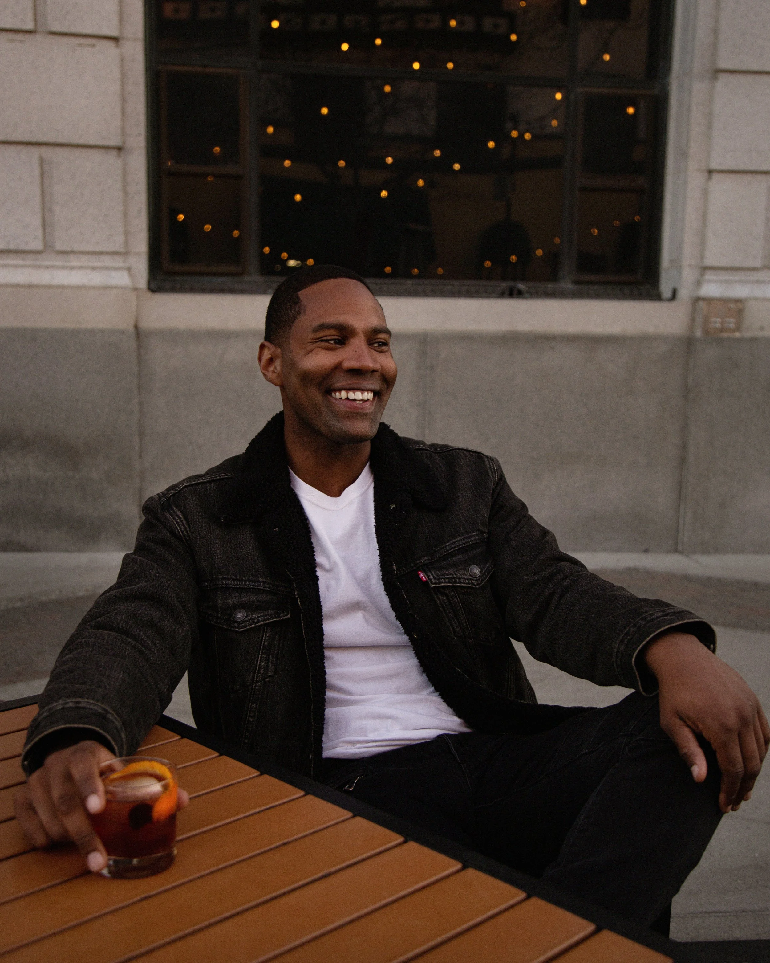 A man sitting at an outdoor table with a drink in his hand, smiling, wearing a black jacket and a white T-shirt, with a window and string lights in the background.