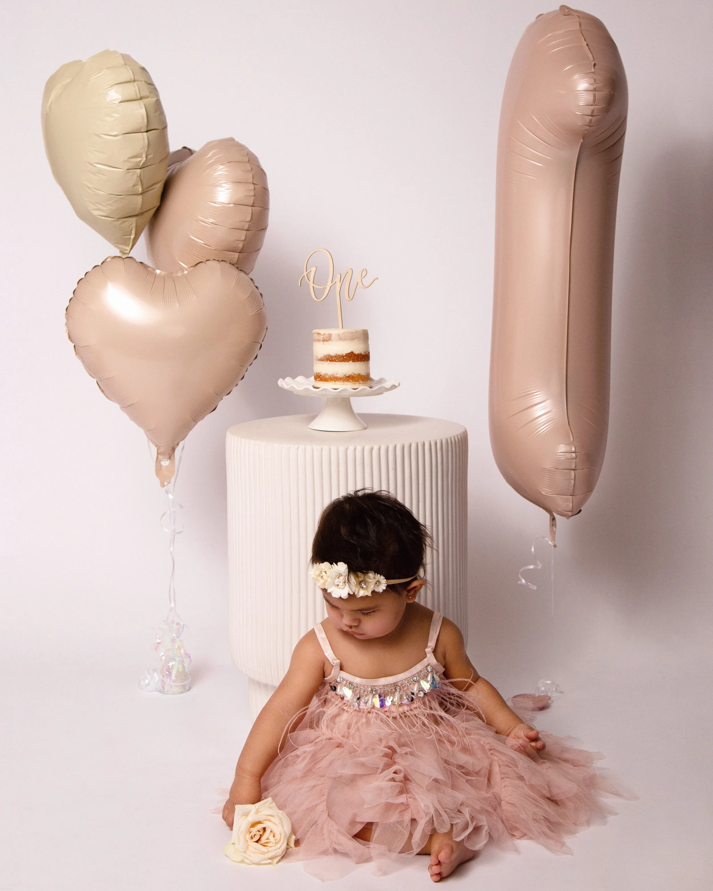 A young girl in a pink tutu dress and floral headband sitting on the floor holding a large artificial rose. Behind her is a white pedestal with a small birthday cake topped with a cake topper that reads 'One'. To the left are four beige and blush-col
