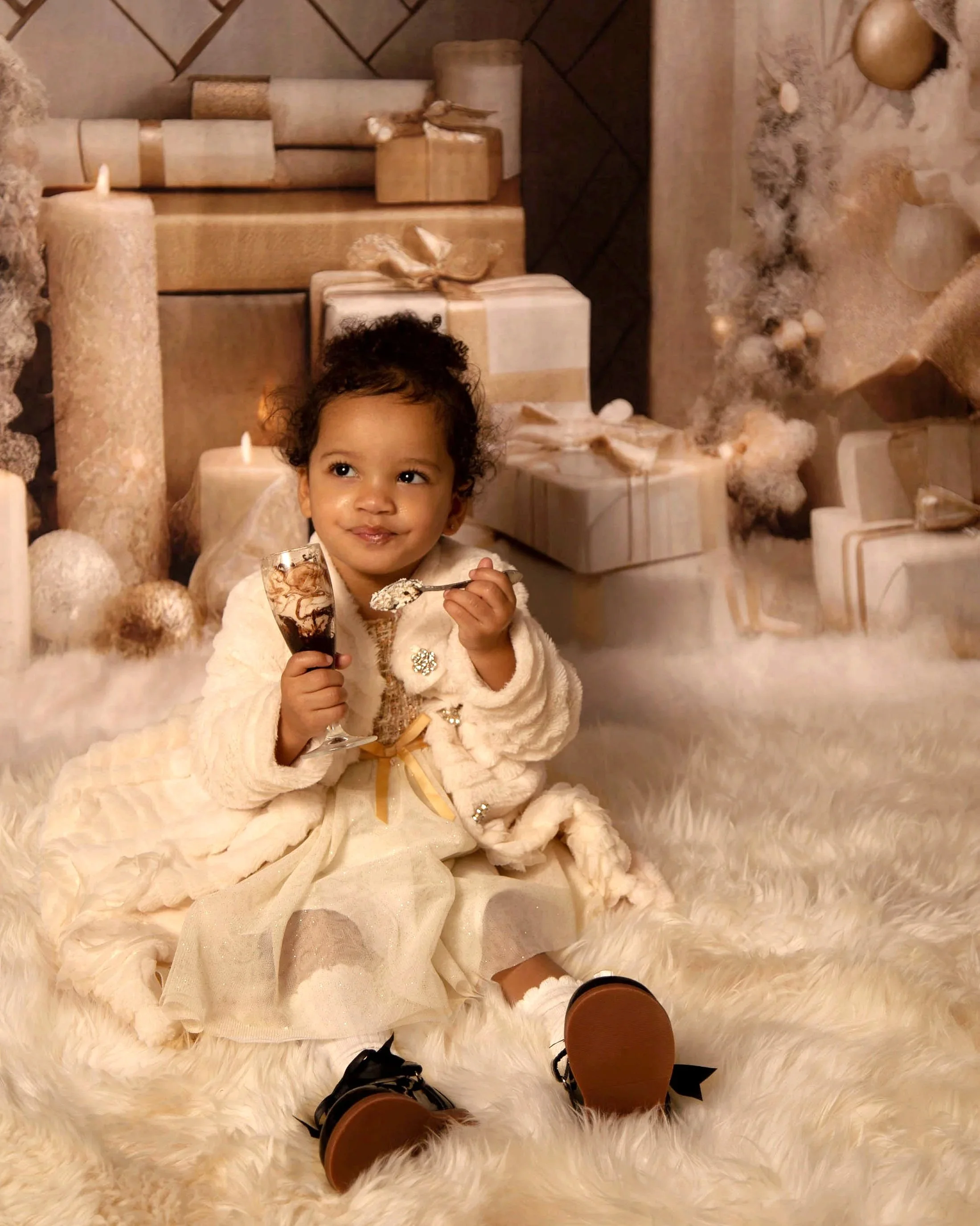 A young girl with curly hair sitting on a fluffy rug, holding a spoonful of ice cream and a glass of chocolate milk, in front of a festive background with white and gold wrapped presents, candles, and holiday decorations.