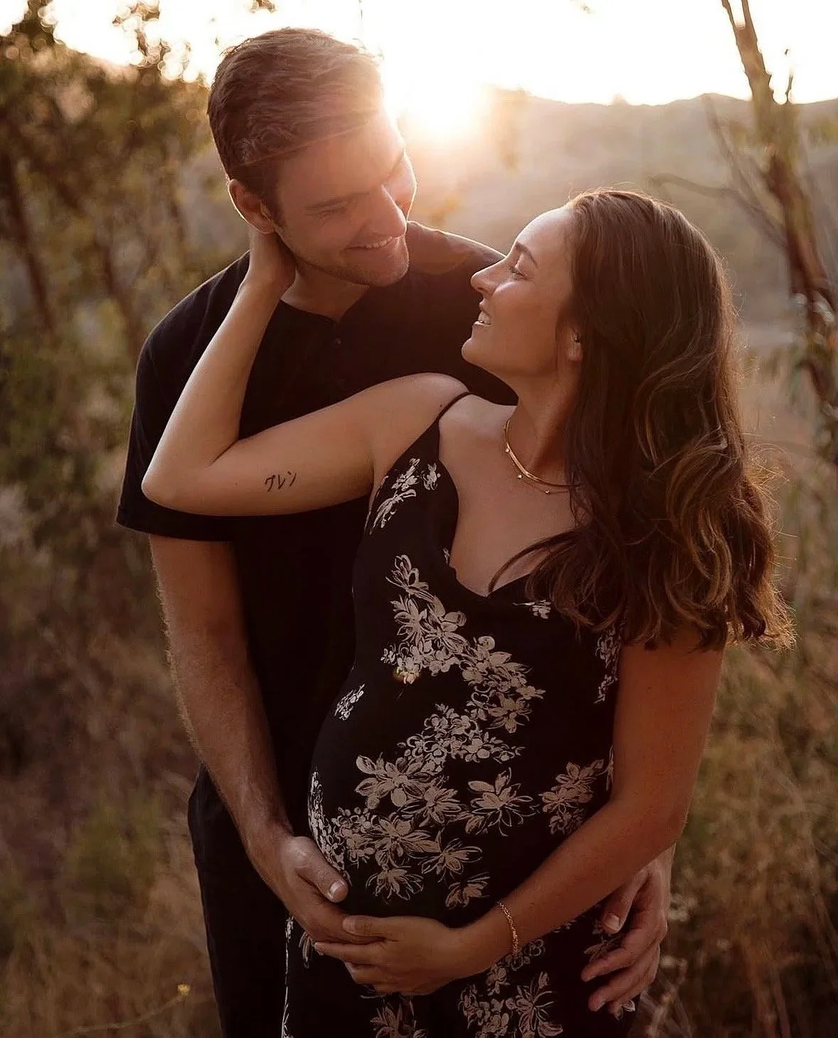 A couple standing outdoors during sunset, with the man embracing a pregnant woman, smiling and looking at each other lovingly.
