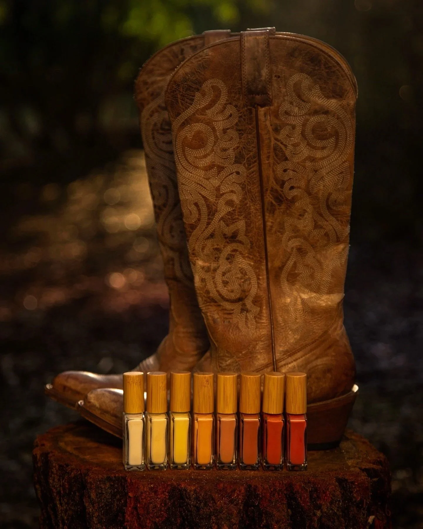 A pair of brown leather cowboy boots with intricate designs, placed on a tree stump, with six small bottles of nail polish in gradient shades from light to dark in front.