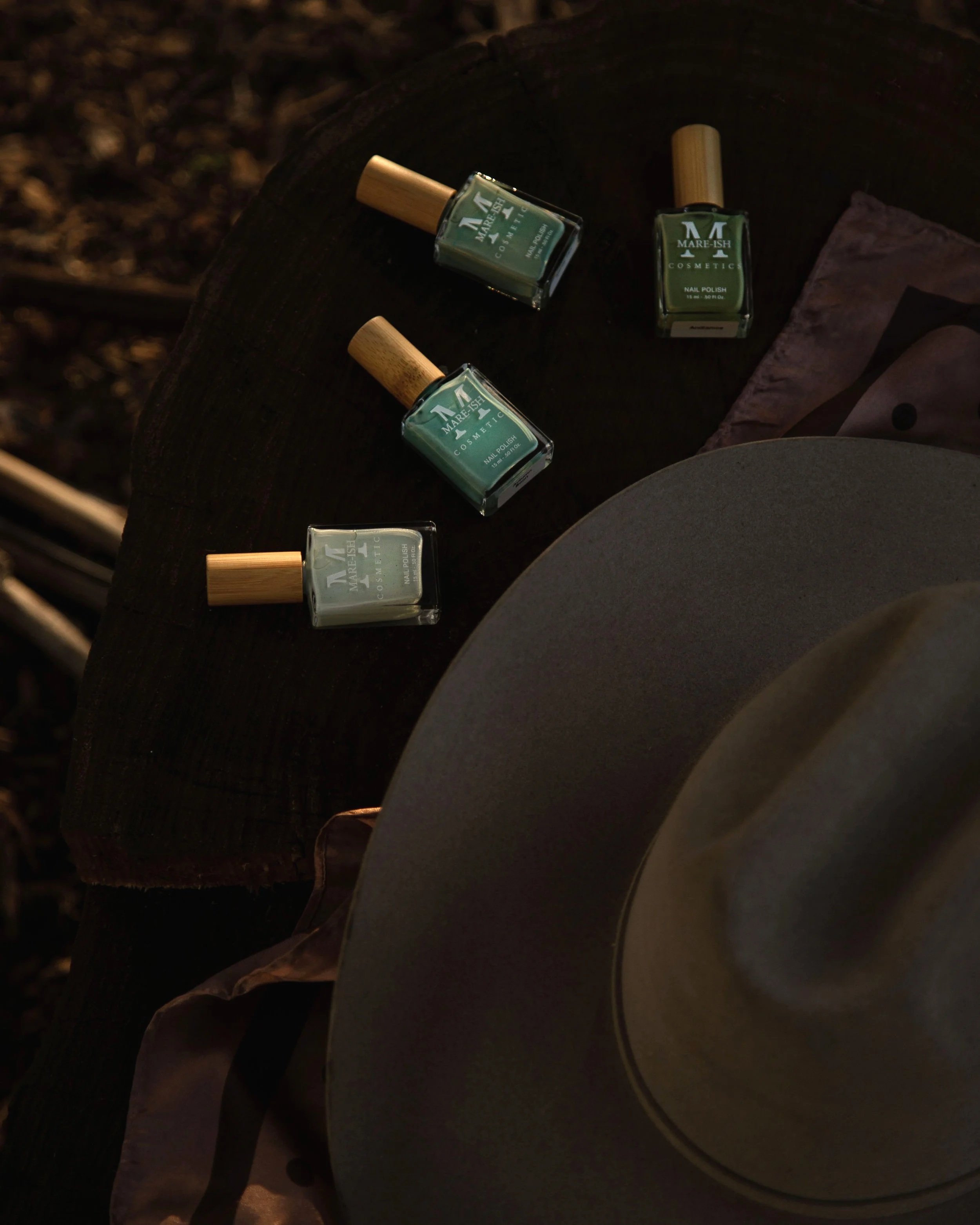 Four bottles of MARE-ISH cosmetic nail polish on a dark wooden surface, along with a beige hat and a piece of cloth in a natural outdoor setting.