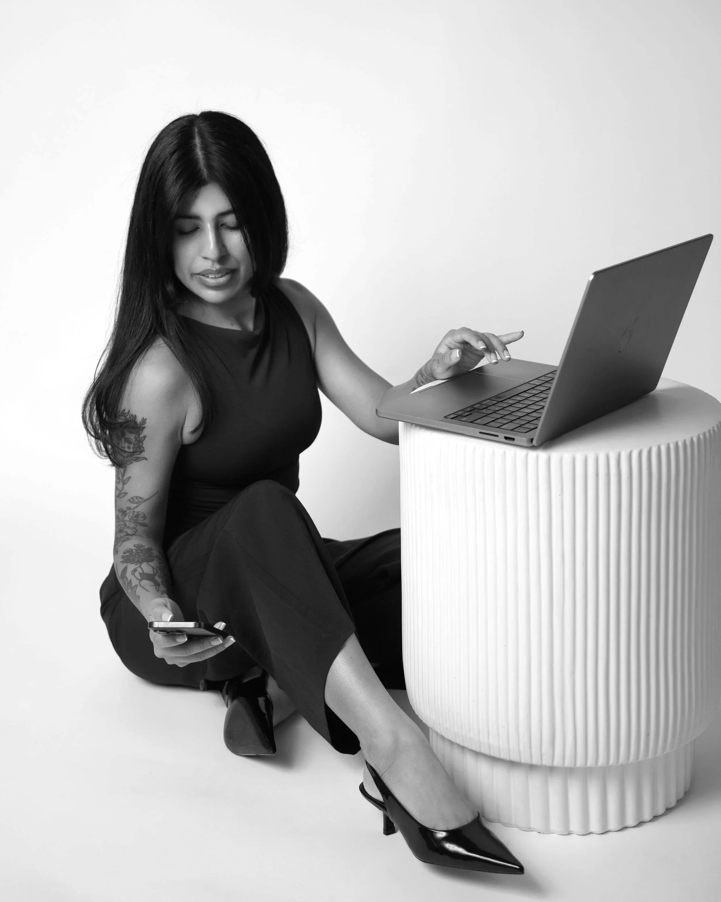 A woman with long dark hair, wearing a sleeveless black top and black pants, sitting on the floor with one leg crossed. She is holding a smartphone in her right hand and using a laptop placed on a round, ribbed white table or pedestal. The background