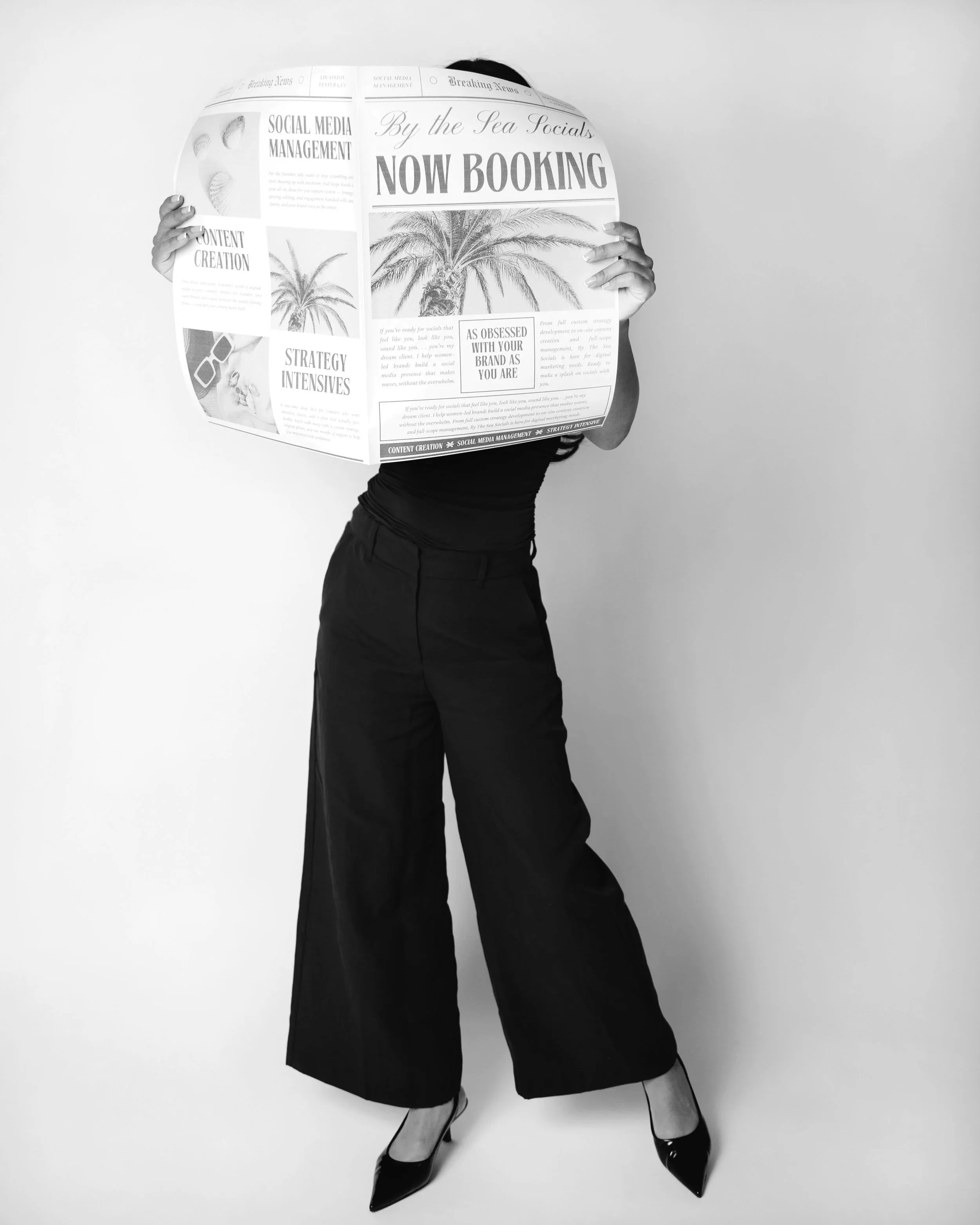 Person standing in front of a plain wall, holding a large newspaper in front of their face, dressed in black pants, high heels, and a black top.
