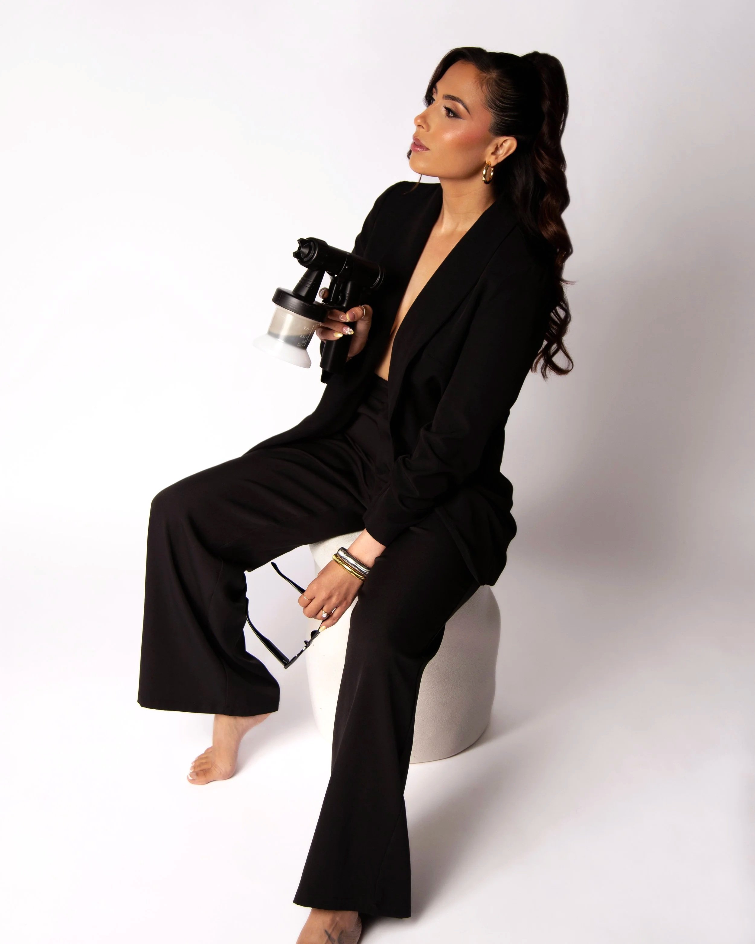 A woman dressed in a black blazer and pants sitting on a small white stool, holding a spray paint machine, with long dark hair styled in waves against a plain white background.