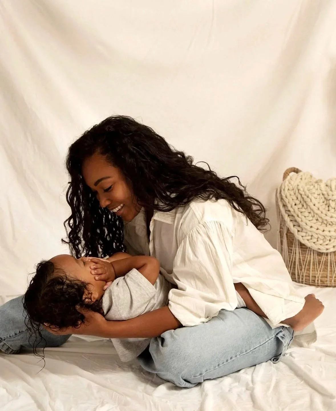 A woman and a young boy playing on a bed, with the woman lifting the boy in her arms. The background is plain with a knitted basket.