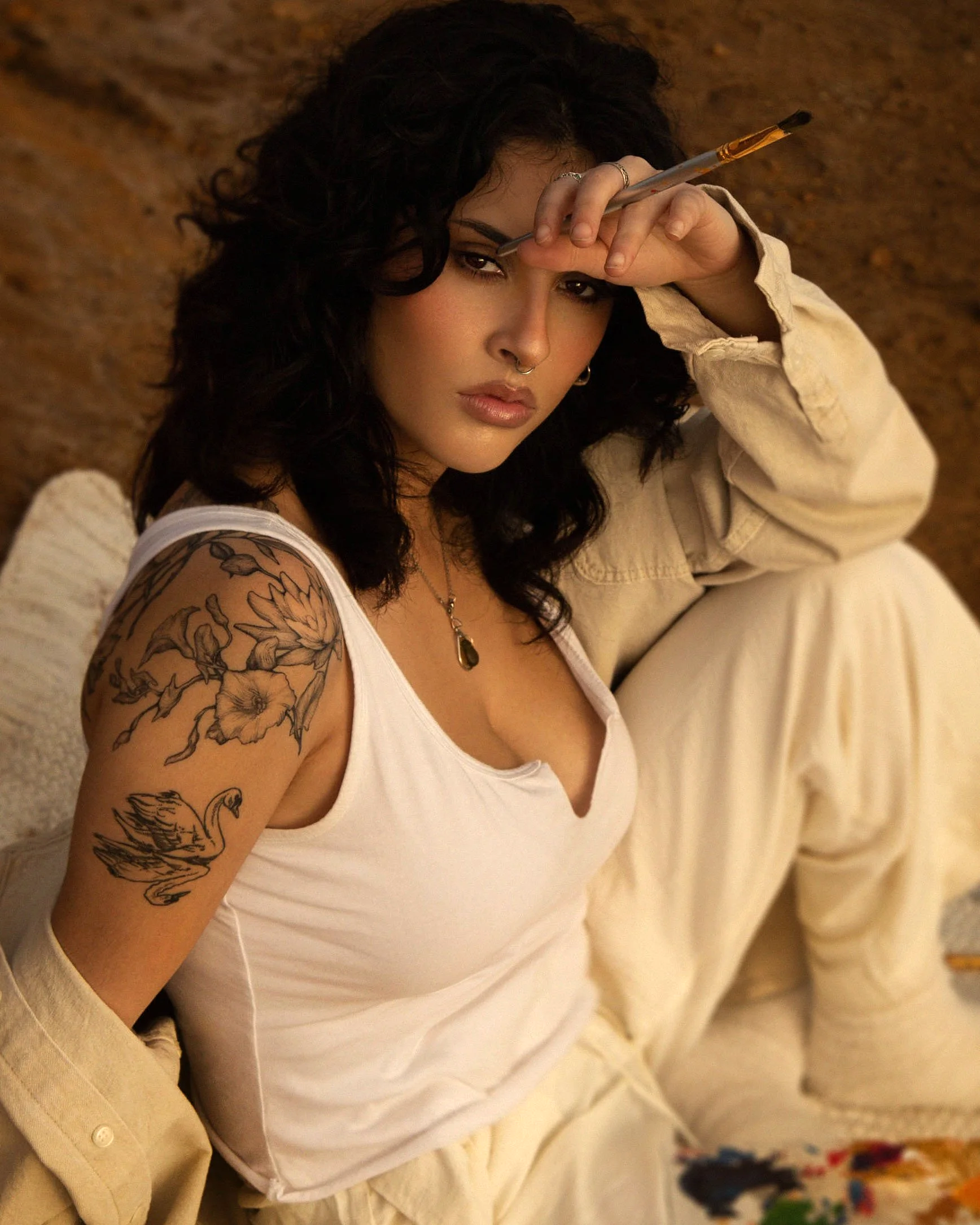 A woman with dark curly hair, tattoos on her left arm, and jewelry holding a paintbrush to her forehead while sitting against a textured wall.