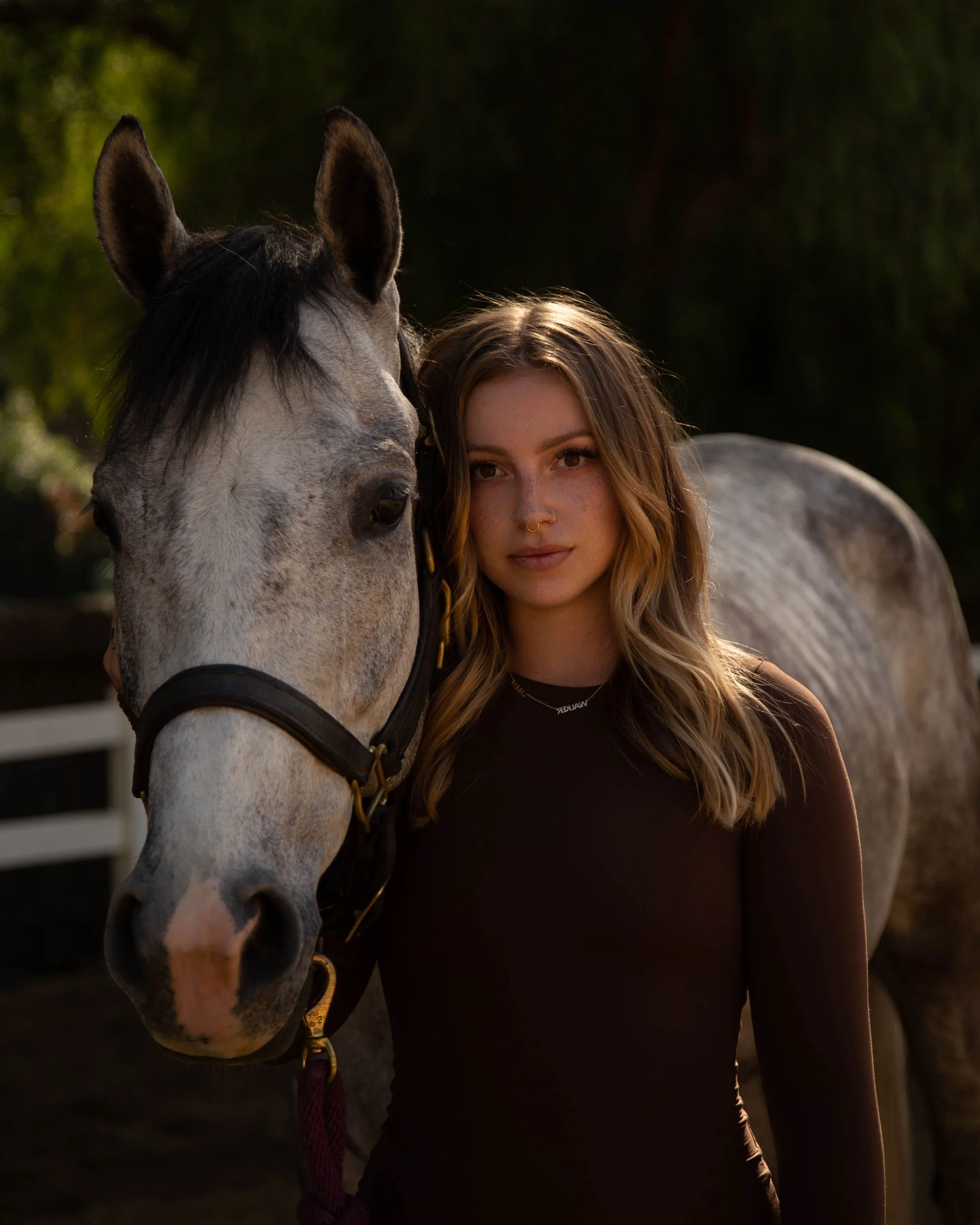 A young woman with wavy blonde hair and a septum piercing standing next to a gray horse outdoors at sunset.
