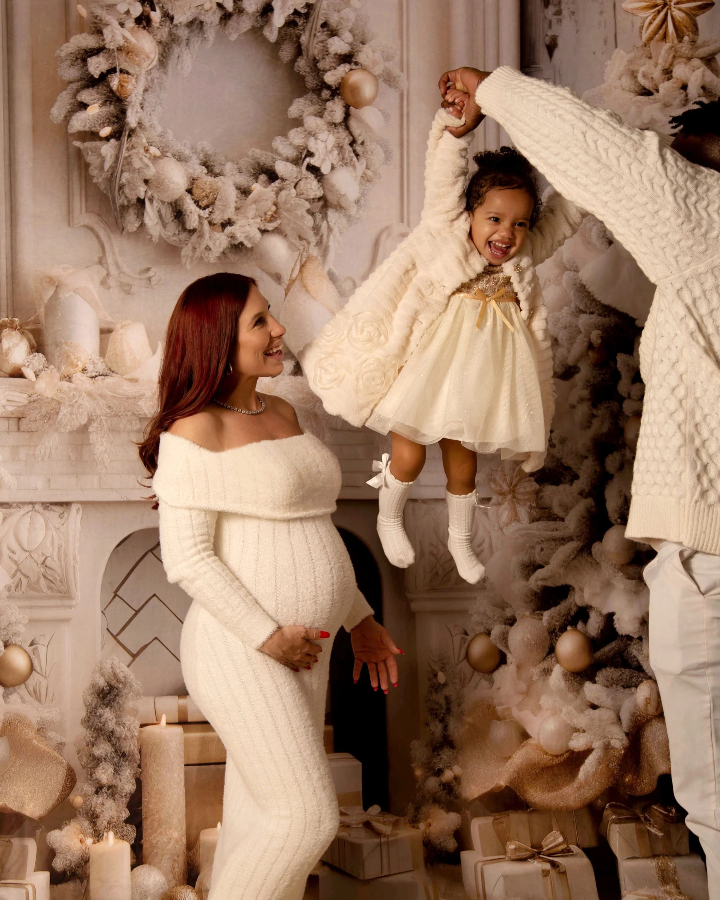 A mother and father with a young girl celebrate Christmas in a decorated room. The girl is being lifted by the father, and the mother watches happily. The scene features a decorated Christmas tree, fireplace, candles, and wrapped presents.