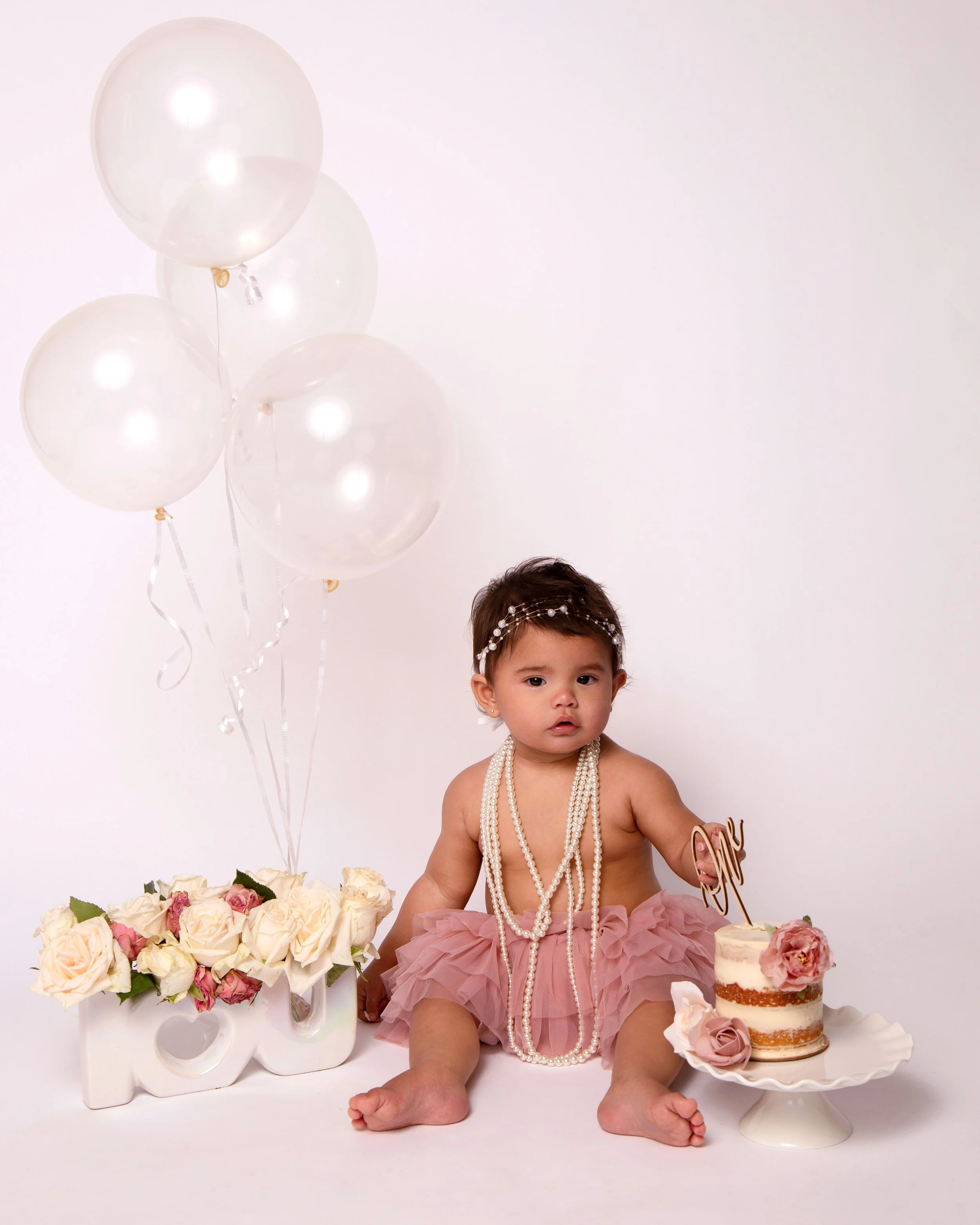 A young girl sitting on the floor during a celebration, wearing pearls and a pink tutu. She is surrounded by white balloons, a pink flower cake with a cake topper that says 'One,' and a decorative white word 'Love' with pink roses.