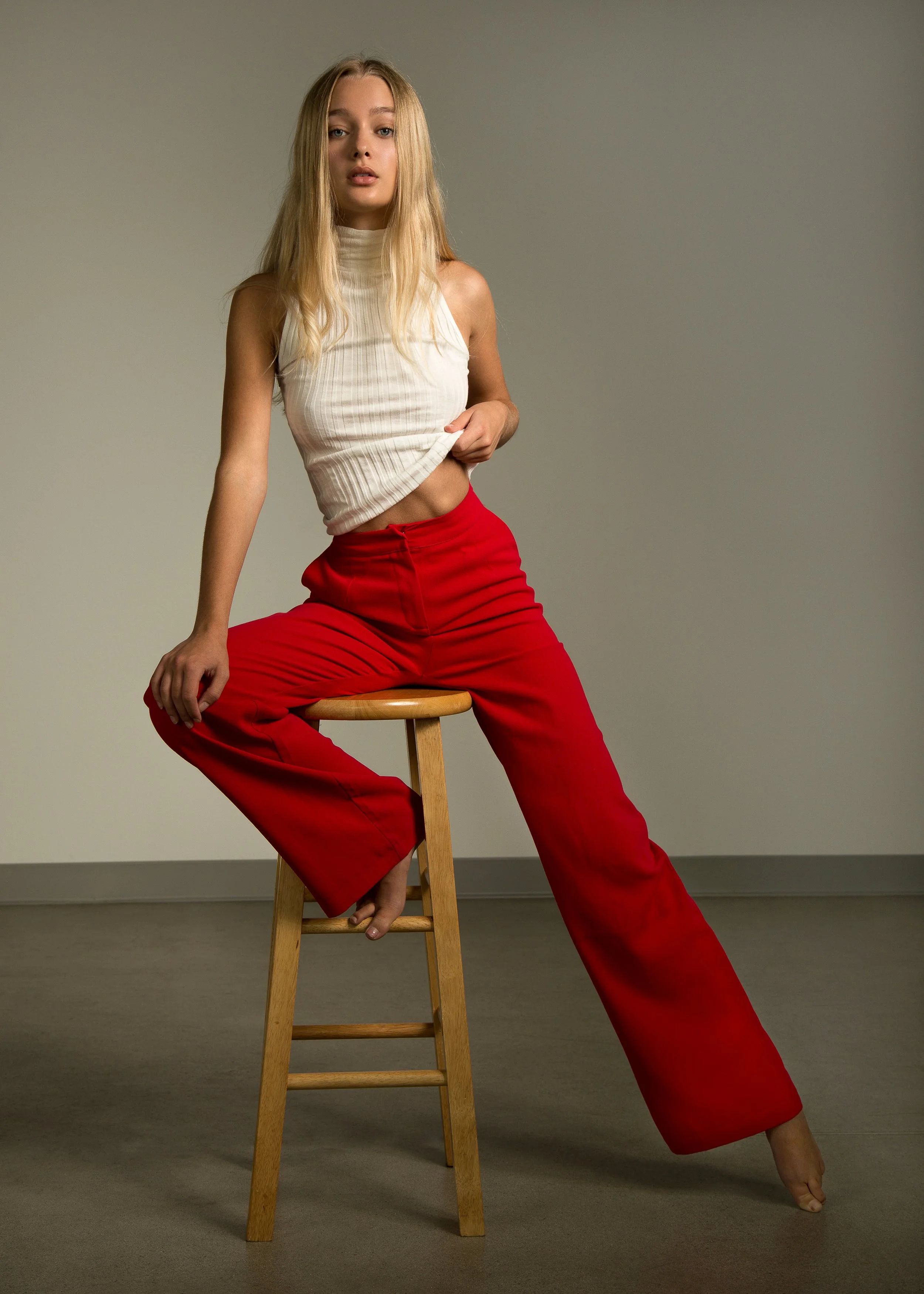 A young woman with long blonde hair sitting on a wooden stool, wearing a sleeveless beige top and red wide-leg pants, against a plain background.