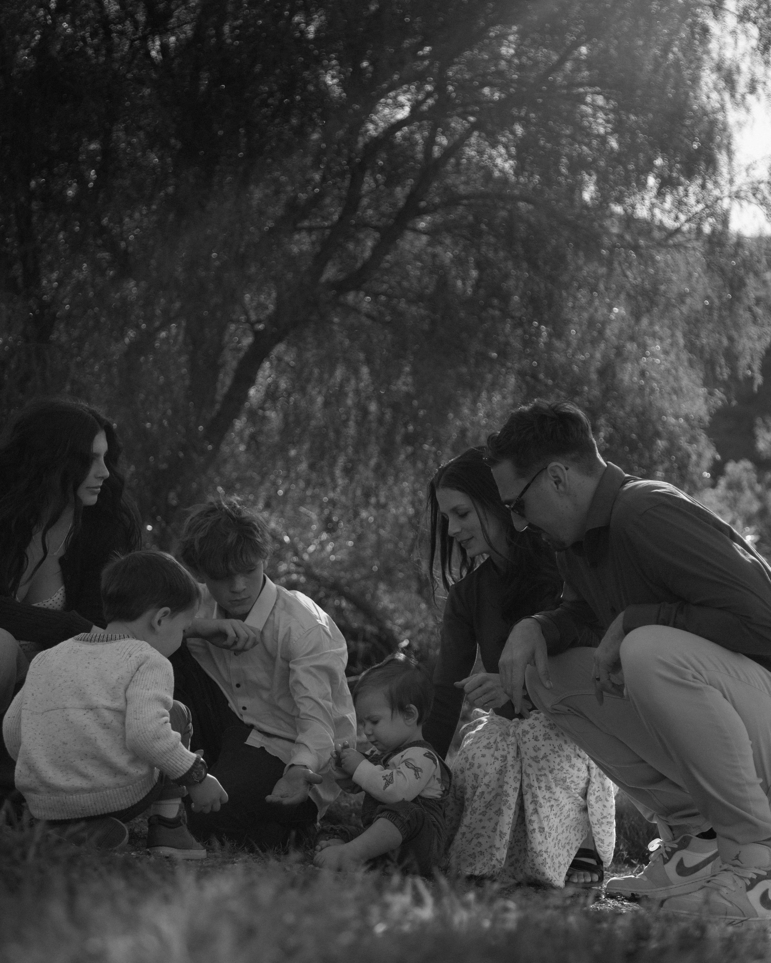 A family of seven, including children and adults, sitting on the grass outdoors under trees, engaging in a shared activity.