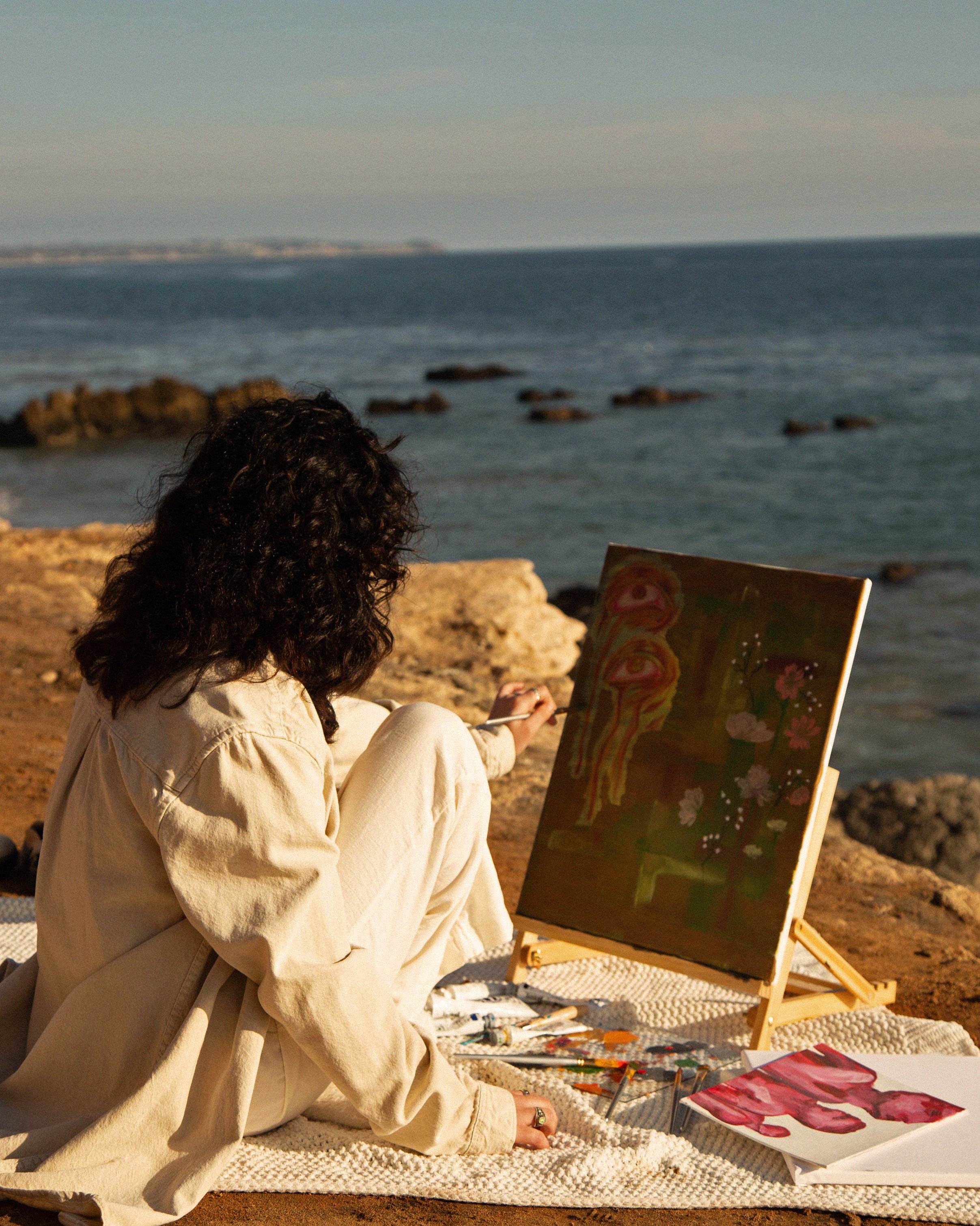 Person with dark curly hair painting on an easel by the water on rocky shore, with painted artwork and art supplies around.