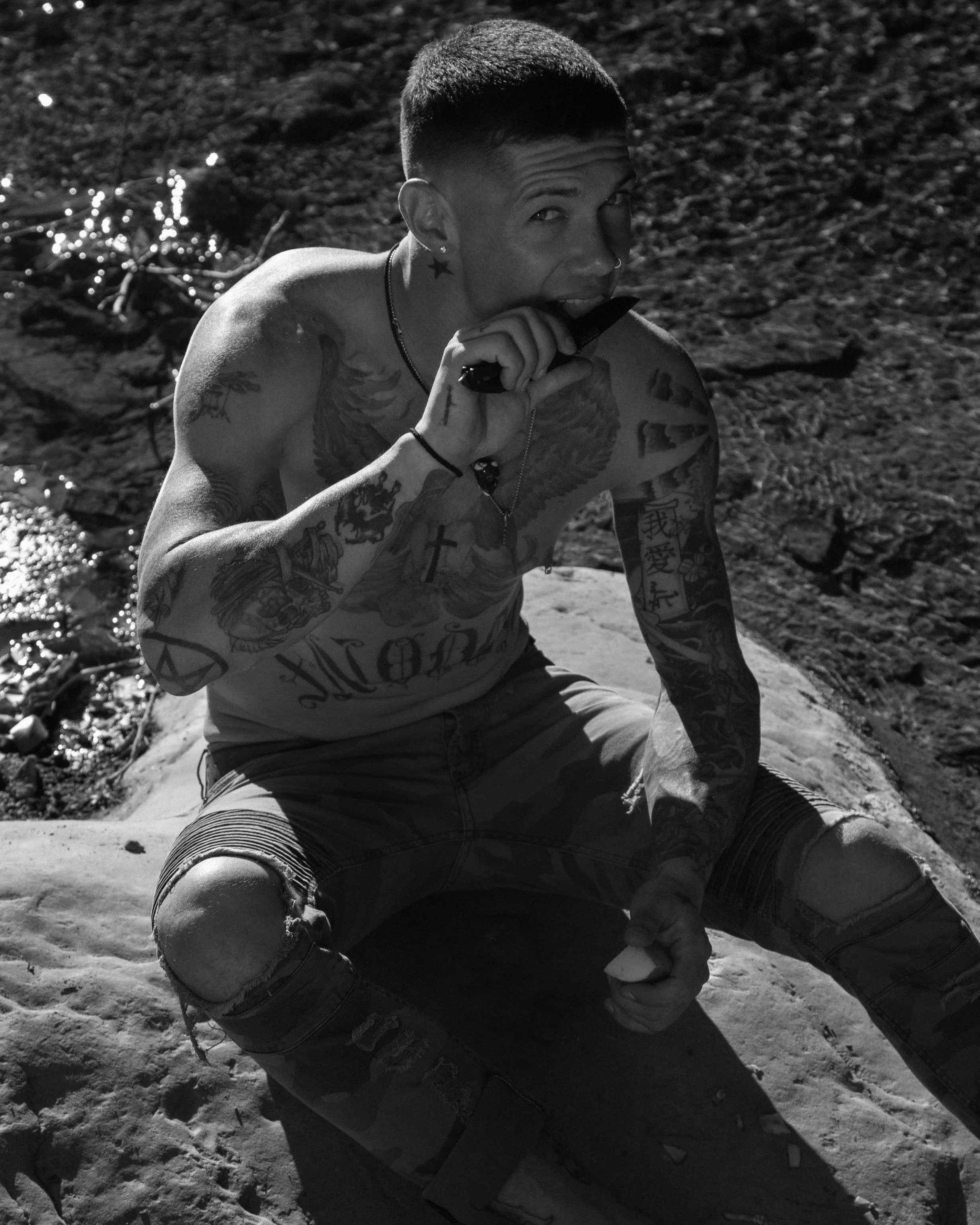 A tattooed man with a buzz cut sitting on a rock by water, holding a knife and biting it, with a smile, in black and white.