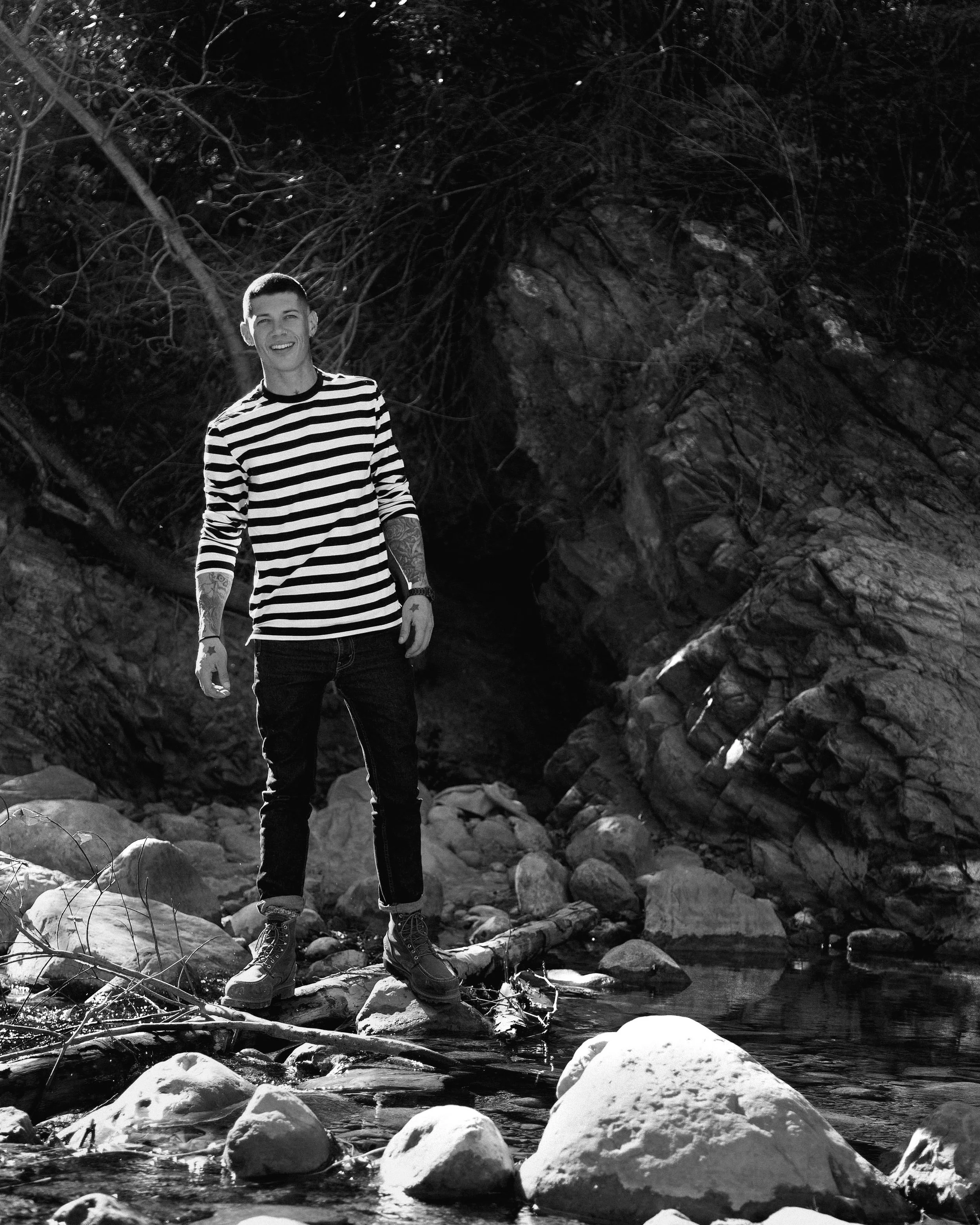 A man standing on rocks in a creek, smiling, with a rocky cave and trees in the background, in black and white.