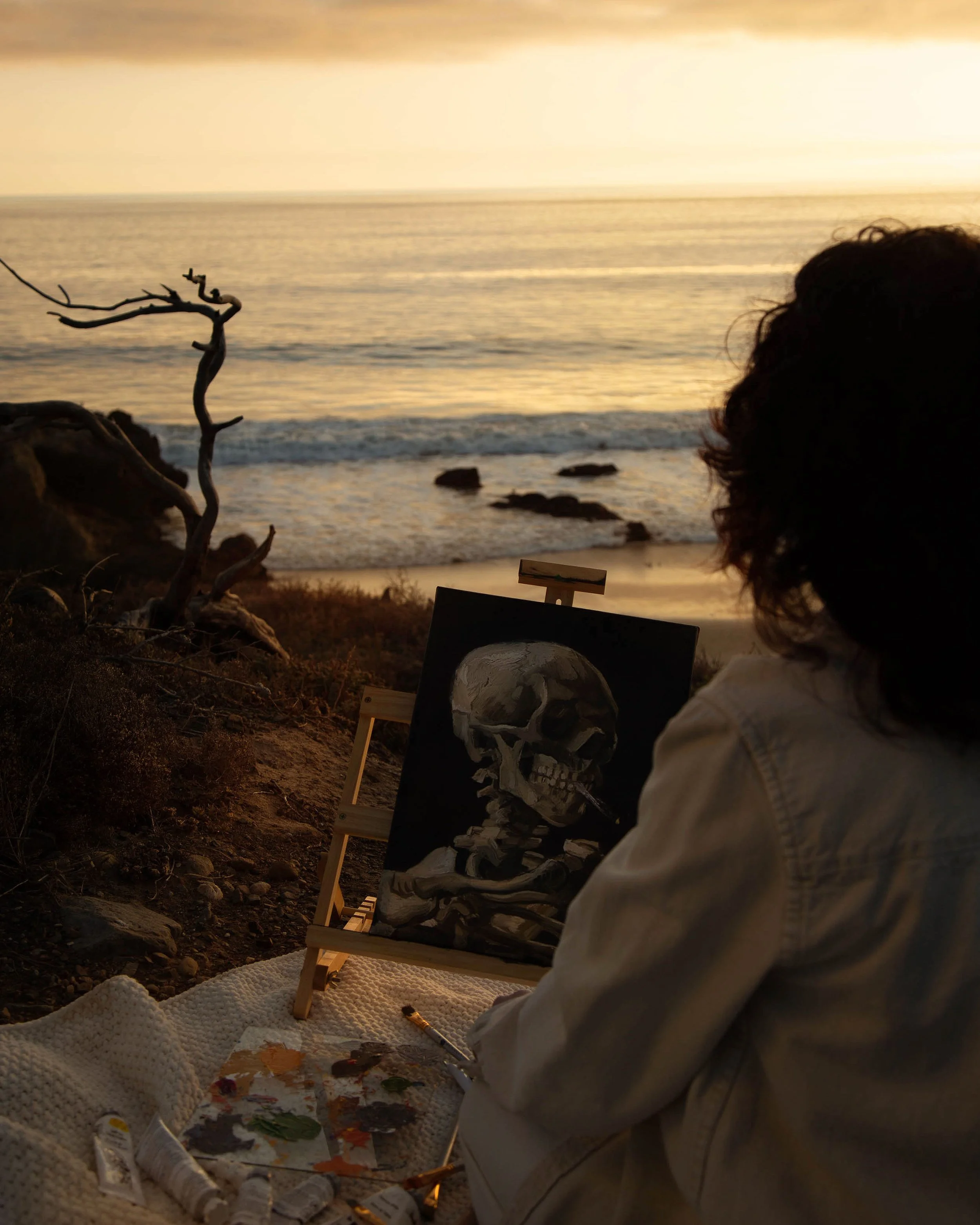 Artist painting a skeleton on a canvas near the ocean at sunset.