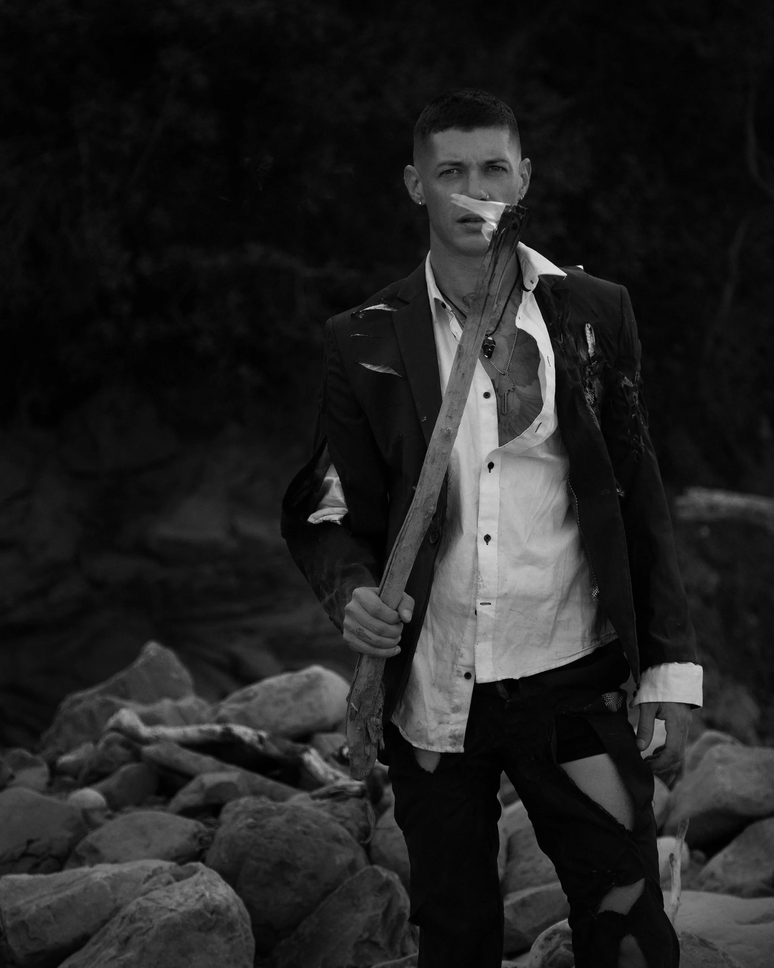 A person with short hair holding a burning torch, standing on rocks in a dark outdoor setting, wearing a torn suit jacket and shirt.