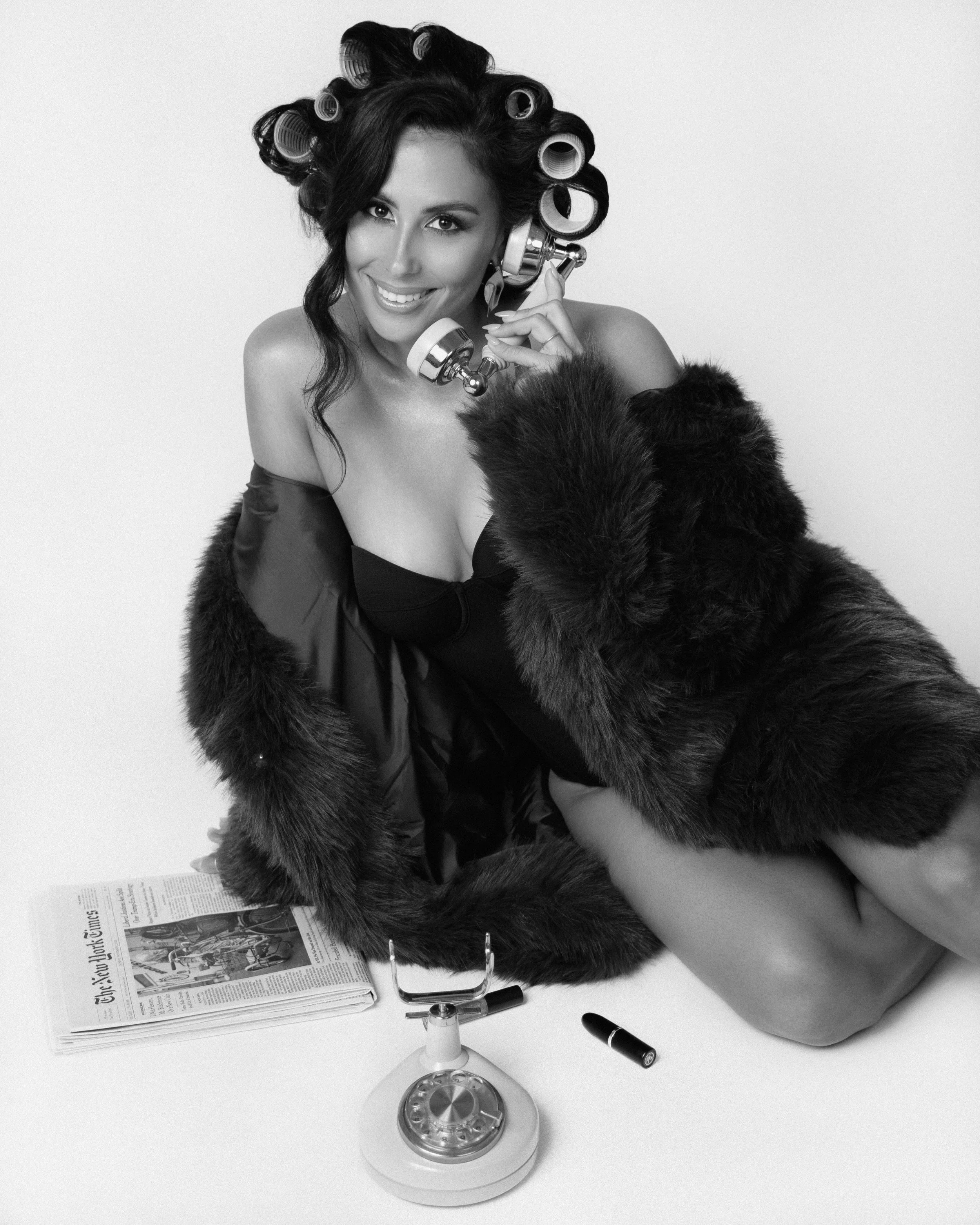 A woman with curlers in her hair talking on a rotary phone, wearing a fur coat and a strapless dress, sitting next to a newspaper, a lipstick, and a microphone.