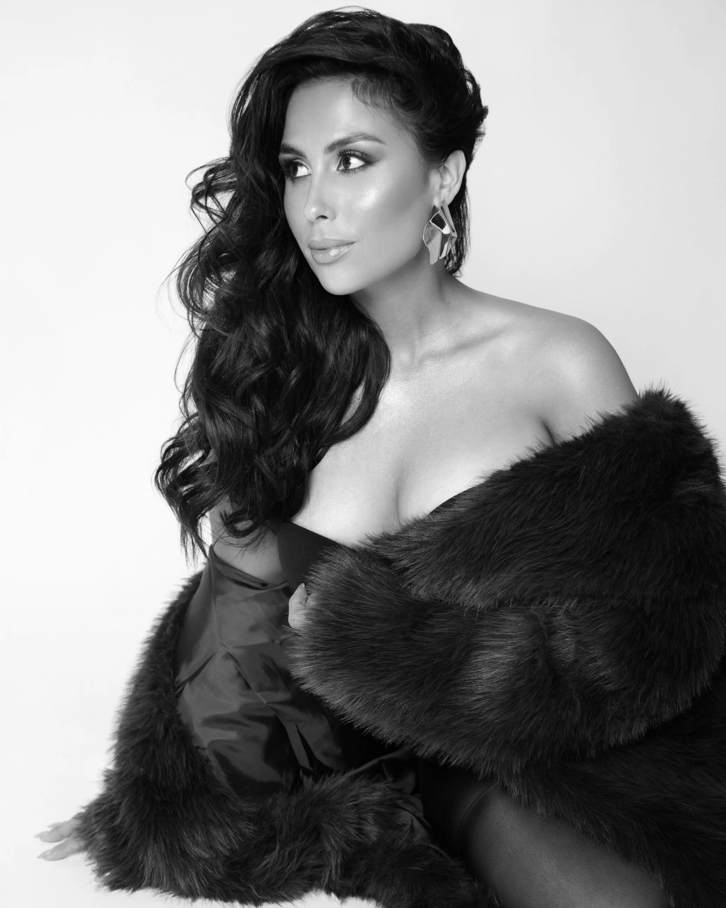 A woman with wavy dark hair, wearing statement earrings and a fur coat draped around her shoulders, looking to the side.