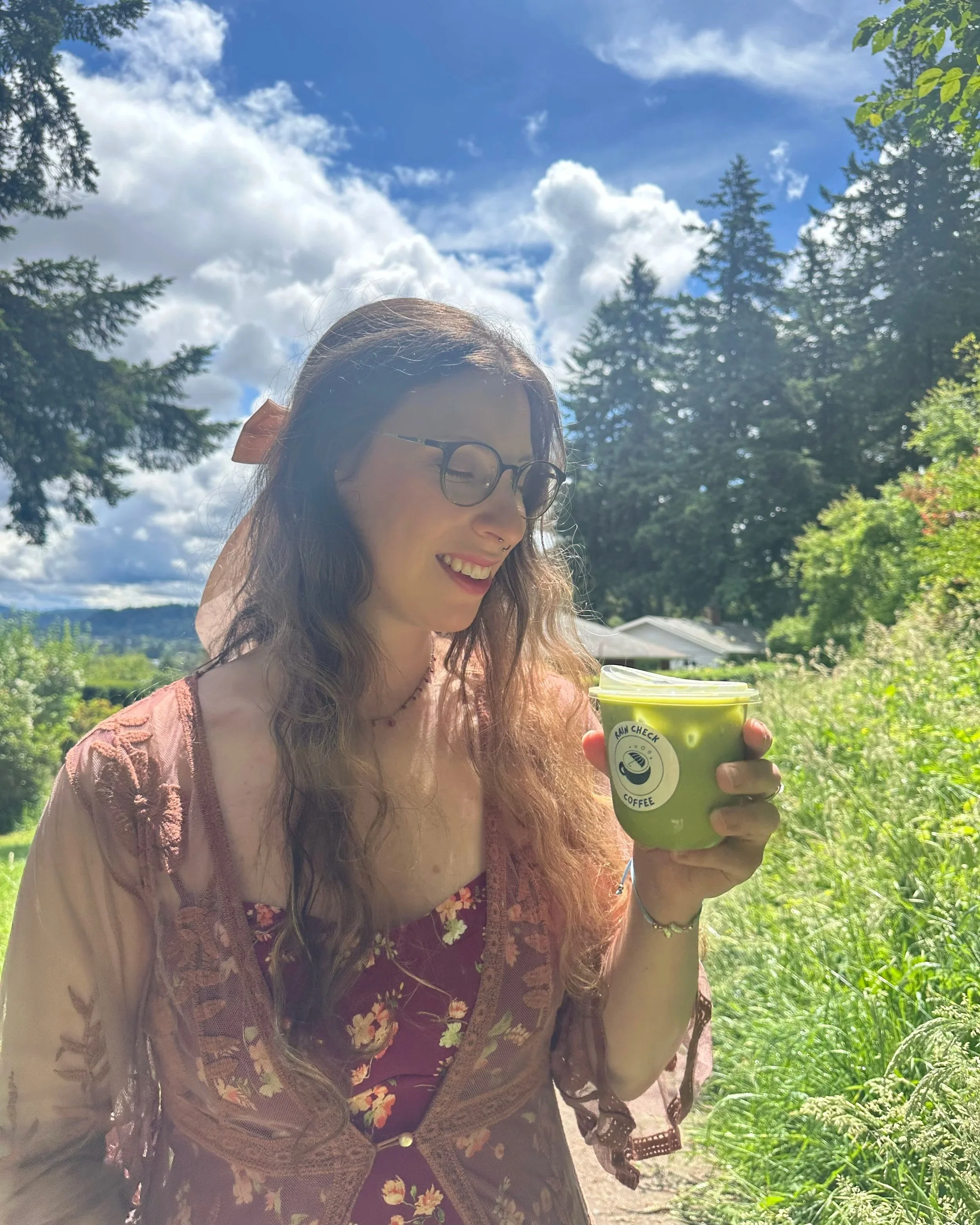 A woman with glasses, long brown hair, and a septum piercing smiling and holding a green cup outdoors on a sunny day, surrounded by trees and a house in the background.