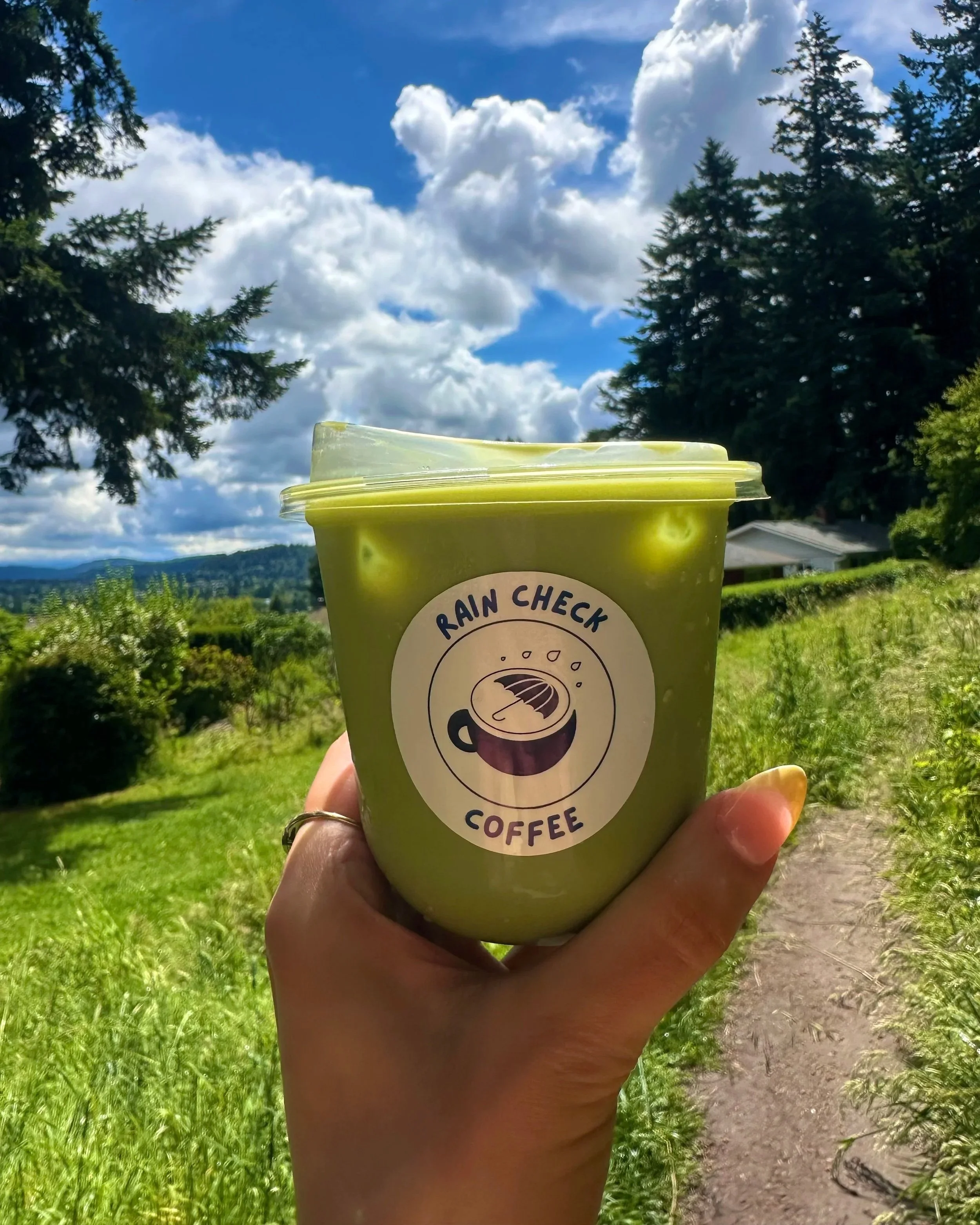 Hand holding a green container labeled 'Rain Check Coffee' against a backdrop of trees, blue sky, and clouds.
