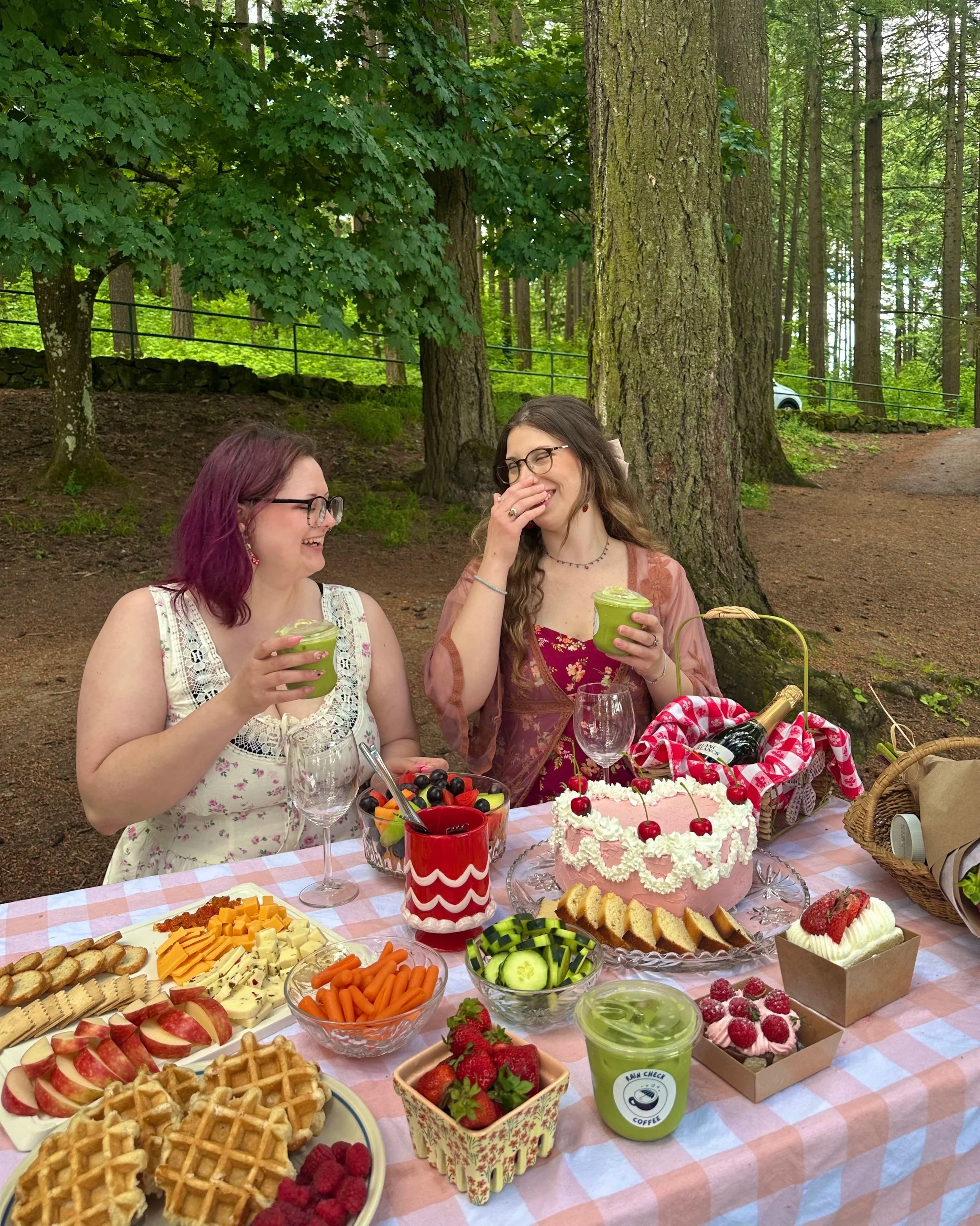 Two women sitting at a table outdoors, laughing and holding green drinks, during a gathering with food including waffles, cheese, fruit, and a pink cake with cherries on top
