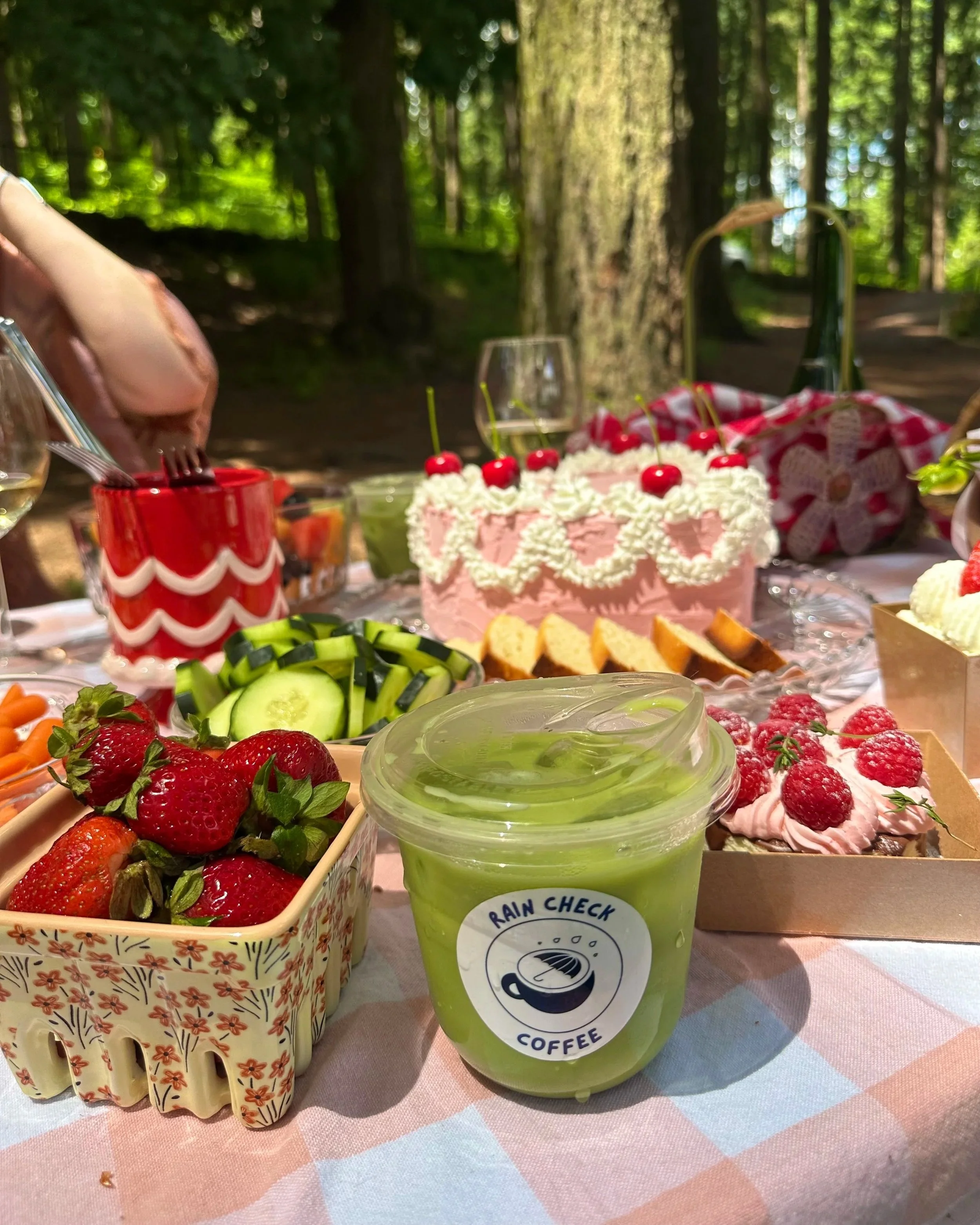 A picnic table set with a variety of food and drinks, including a pink frosted cake with cherries on top, strawberries, sliced cucumbers, and a green smoothie labeled "Rain Check Coffee," in a forested outdoor setting.