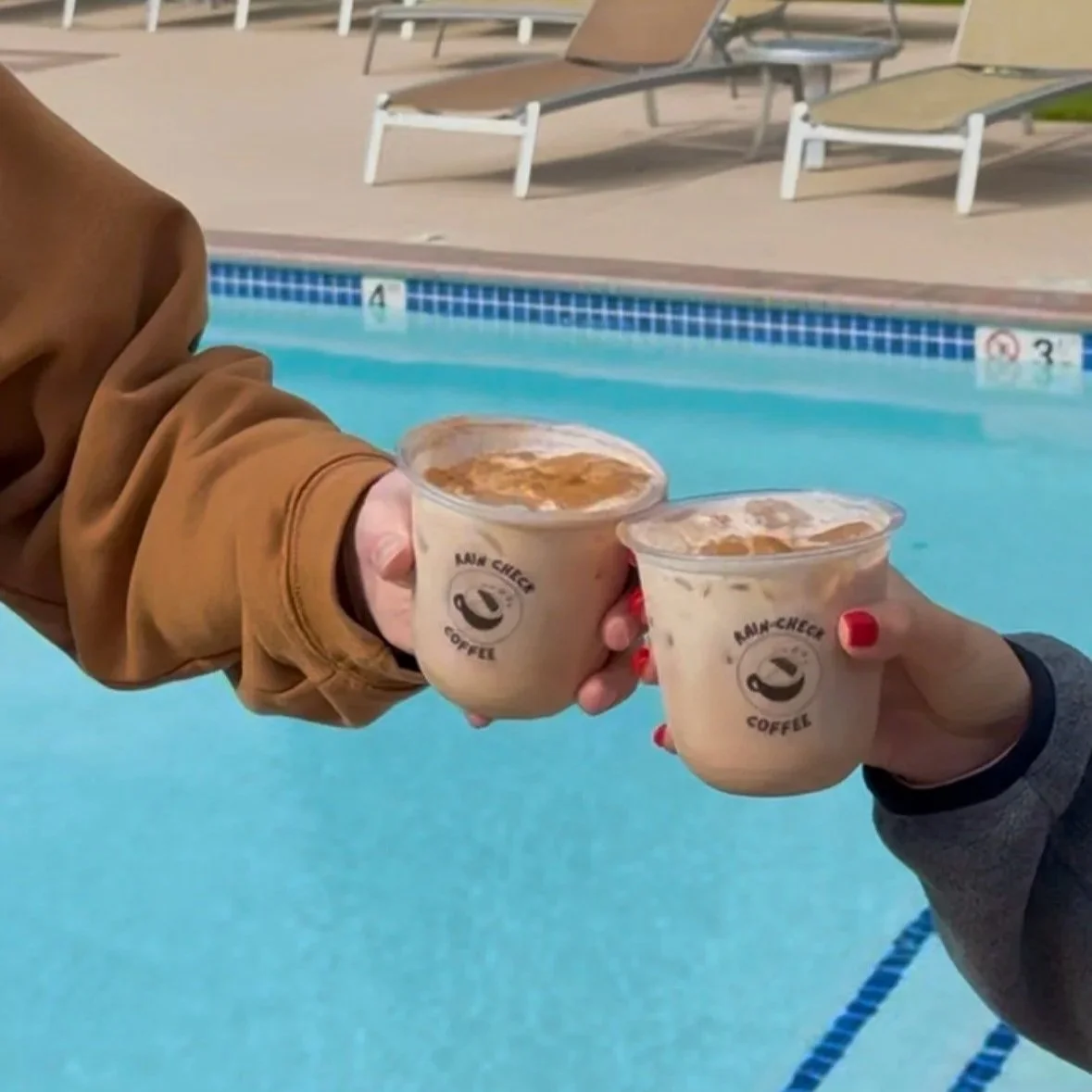 Two people holding cups of iced coffee near a swimming pool, with lounge chairs in the background.