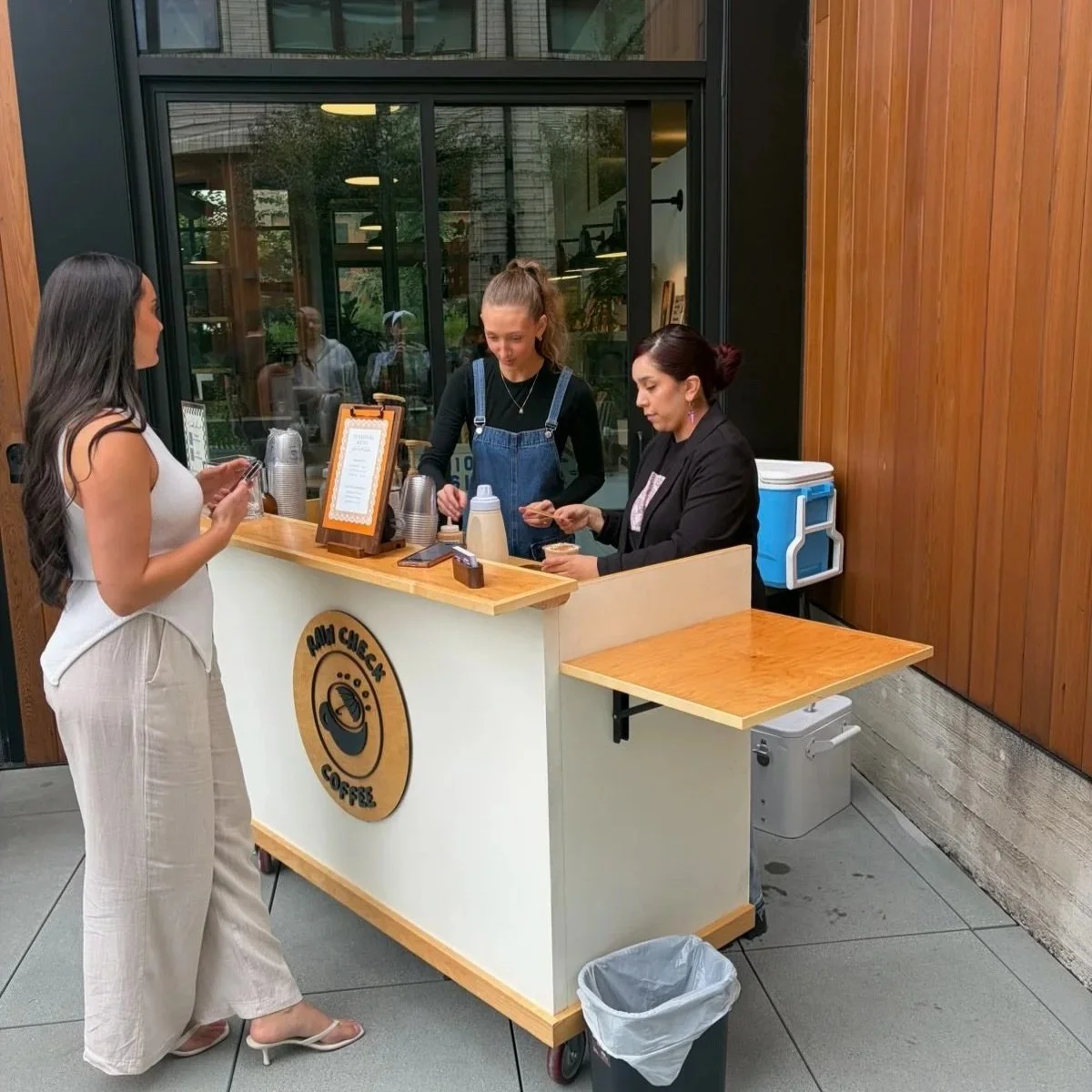 A woman in a white dress placing her order at a coffee stand operated by two baristas, one with a ponytail and the other with dark hair, inside a building with wooden and glass walls.